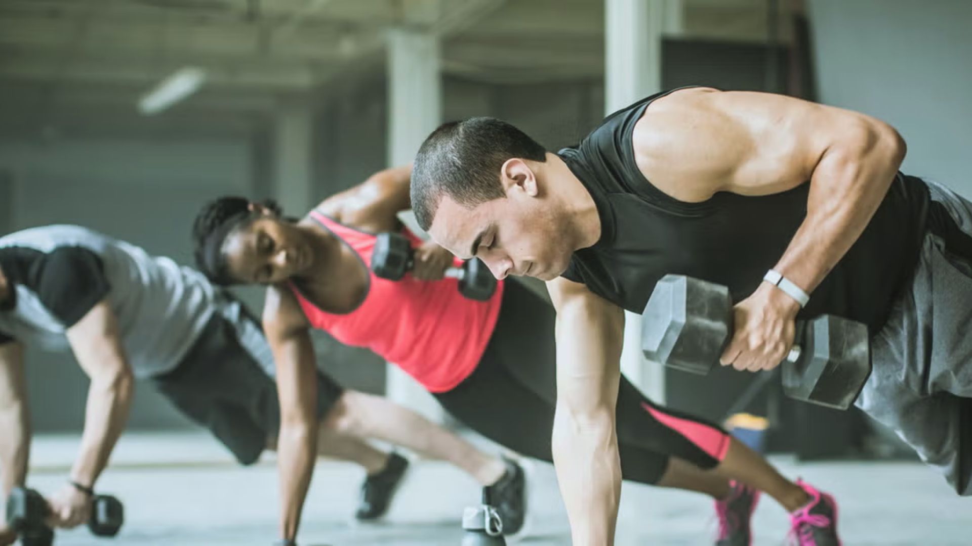 Three people in athletic wear perform dumbbell rows in a gym. They show determination and focus, highlighting strength training in an industrial setting.