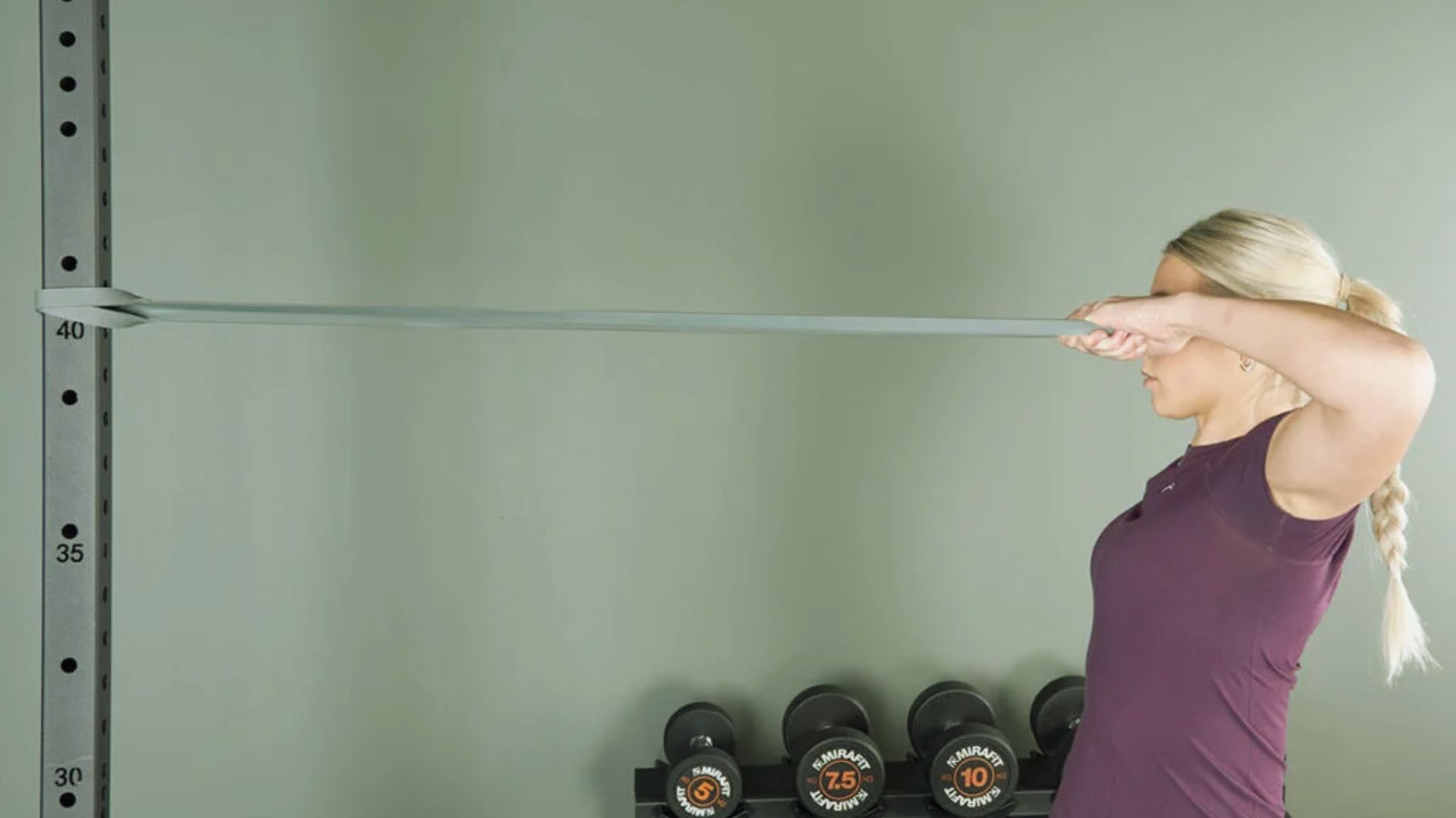 A woman with a blond braid pulls a resistance band at eye level in a gym, facing a rack of dumbbells. She wears a purple shirt, appearing focused and determined.