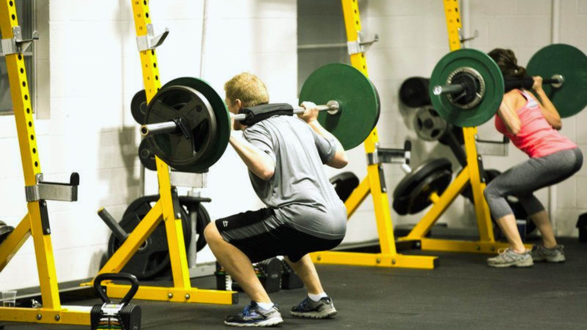 Two people doing barbell squats on a yellow squat rack in a gym, focusing on strength and fitness. The atmosphere is energetic and focused.