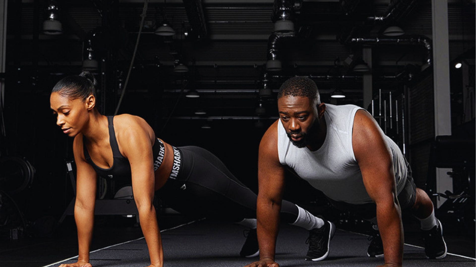 Two individuals performing push-ups in a dimly lit gym. The woman on the left wears a black sports outfit, while the man on the right wears a white tank top. Focused and determined expressions.