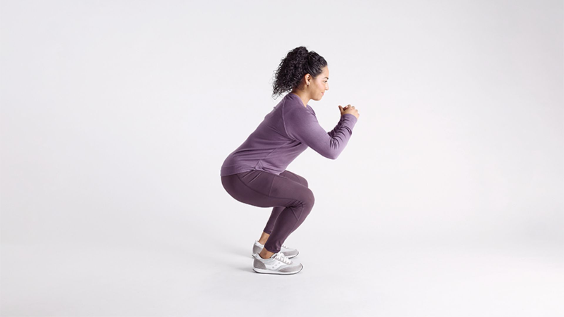 A woman in a lavender workout outfit performs a squat against a plain background. She appears focused and balanced, embodying fitness and strength.