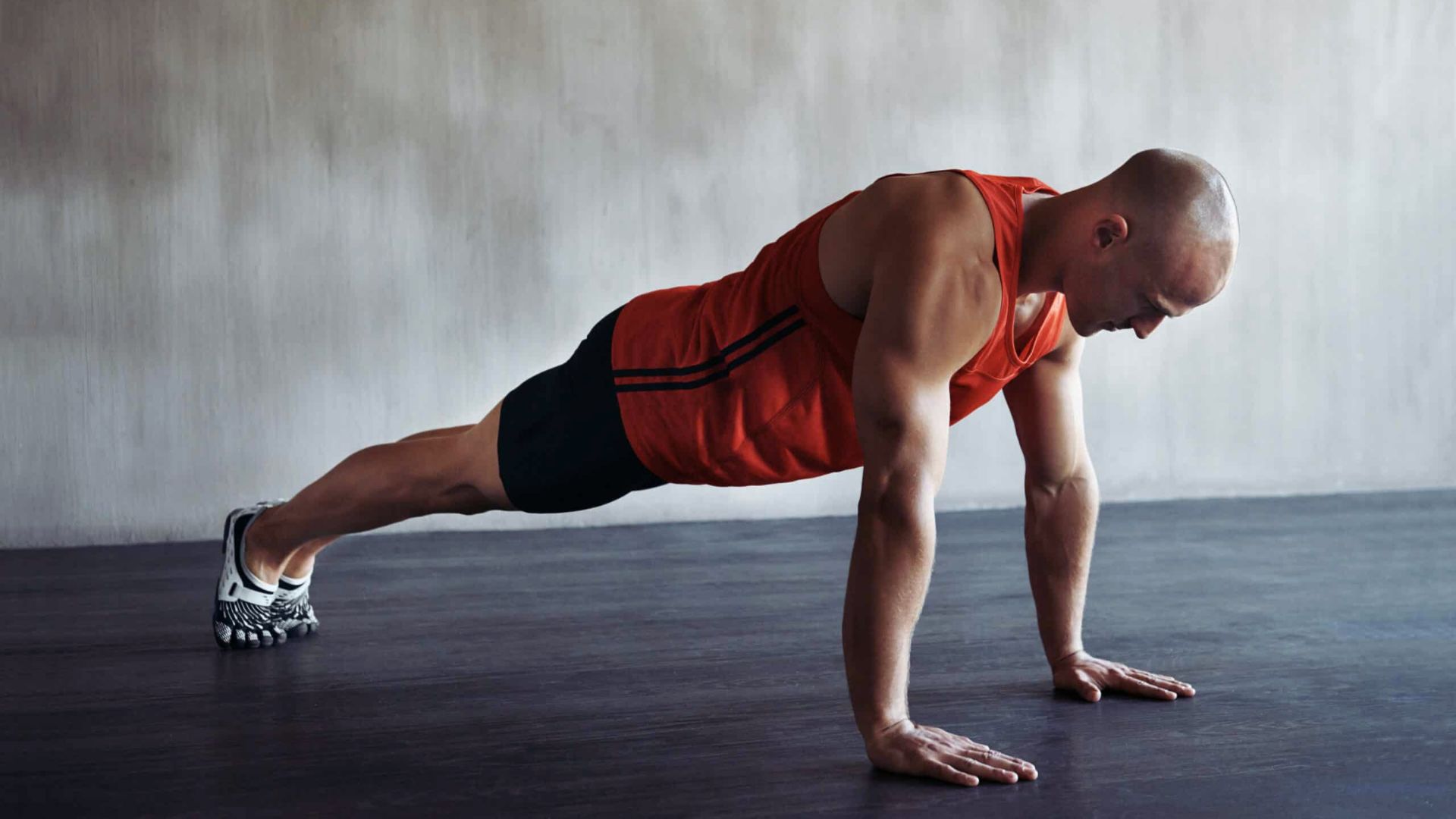 A person in a red tank top and black shorts performs a push-up on a dark wooden floor, exuding focus and strength in a minimalist setting.