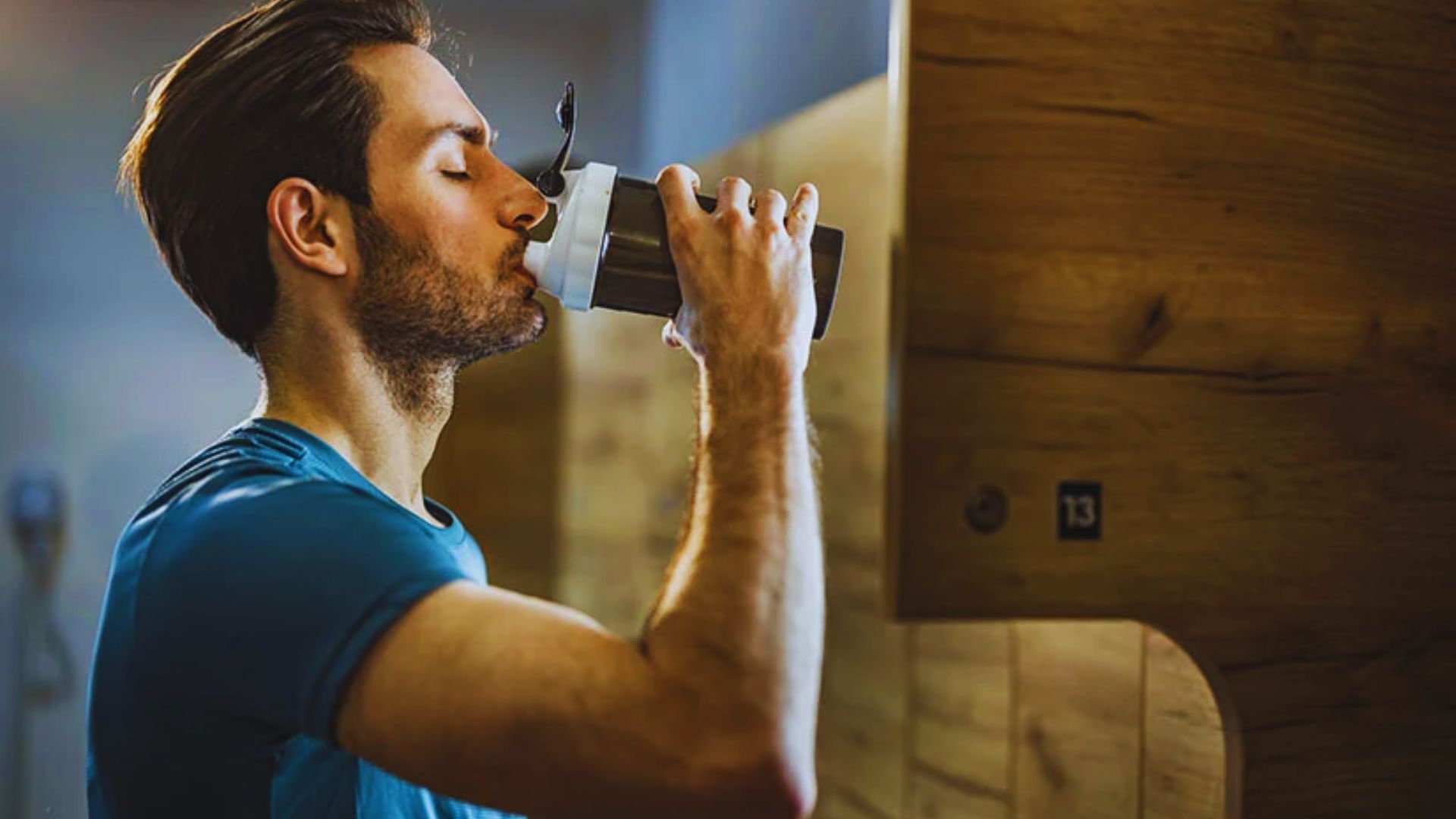 Man in a blue shirt drinks from a protein shaker, post-workout, in a gym locker room. Warm lighting highlights a focused and refreshed expression.