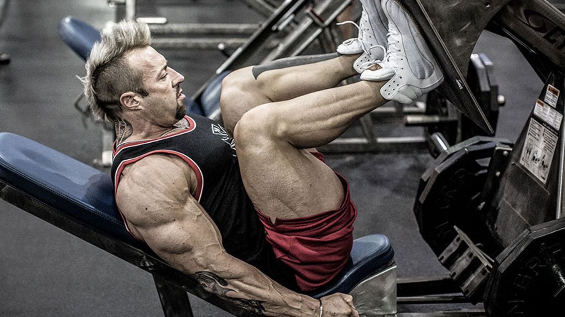 A muscular man with a mohawk hairstyle performs a leg press in a gym, wearing a black tank top and red shorts. The tone is intense and focused.