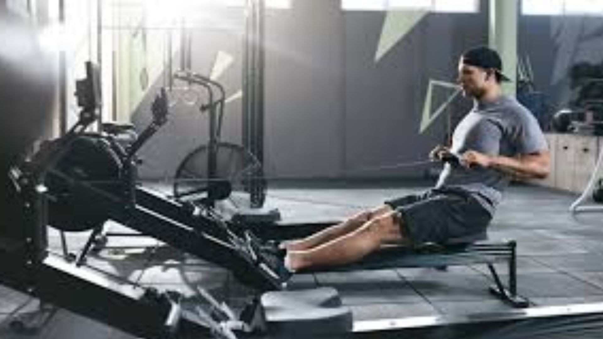 A man in athletic wear uses a rowing machine in a well-lit gym, focused and determined. Sunlight streams in, highlighting gym equipment in the background.