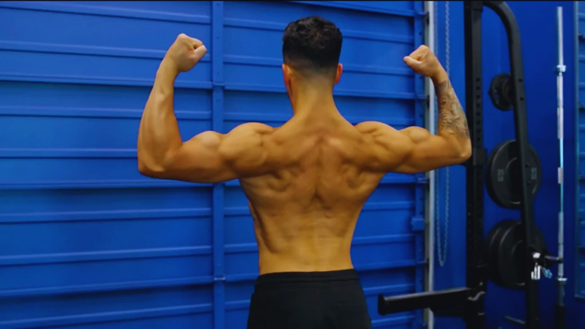 A muscular man facing away flexes his back and arm muscles against a bright blue gym wall, conveying strength and focus.