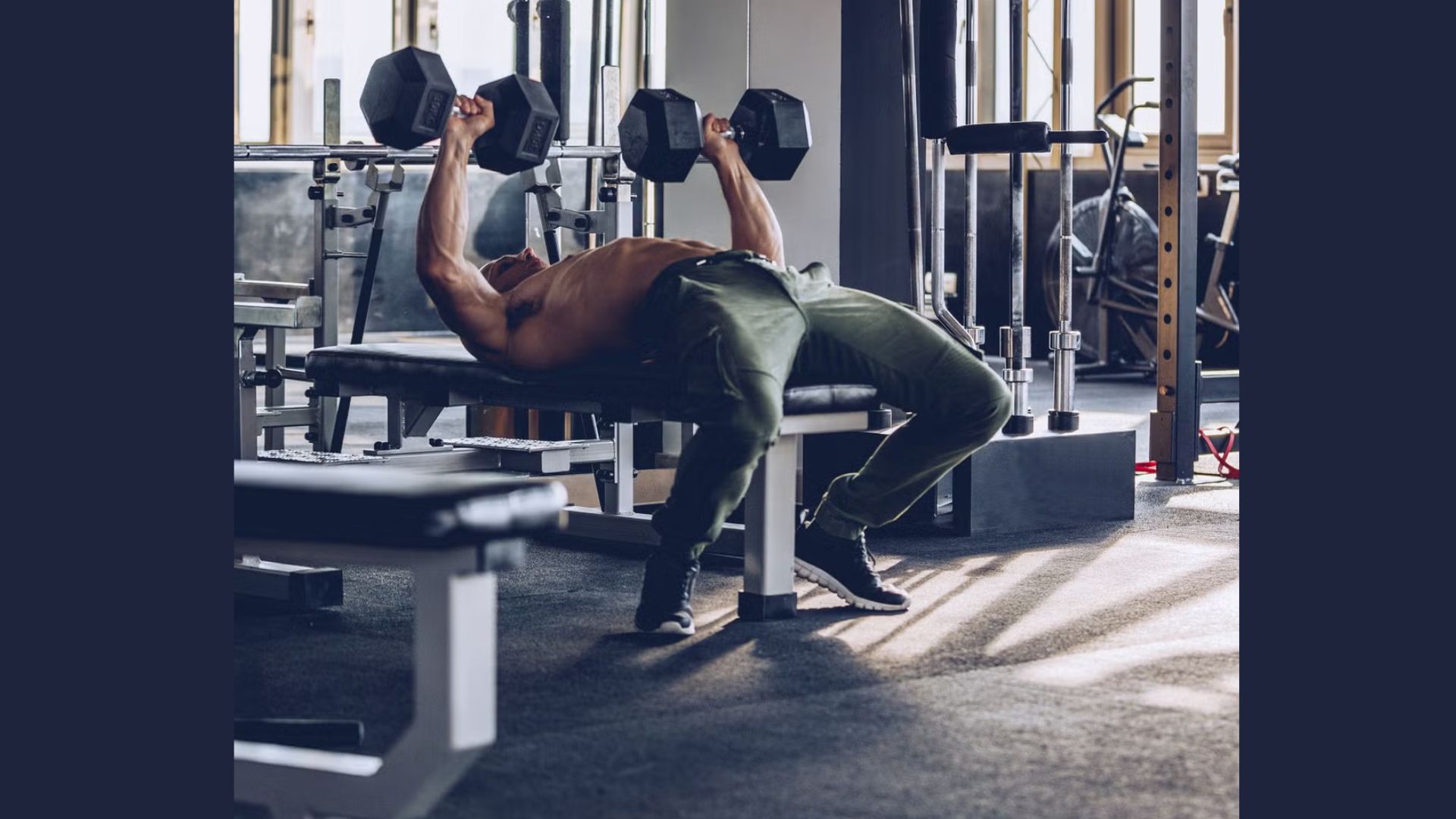 A person in a gym performs a bench press with dumbbells on a bench. The setting is bright and modern, evoking a sense of focus and strength.