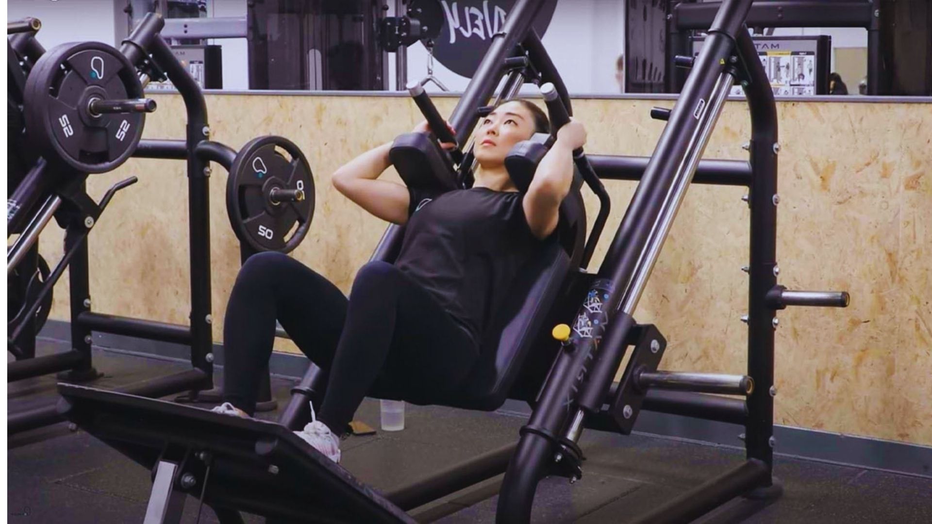 A person is focused and determined while using a leg press machine at a gym. They are wearing a dark outfit, surrounded by weights and gym equipment.