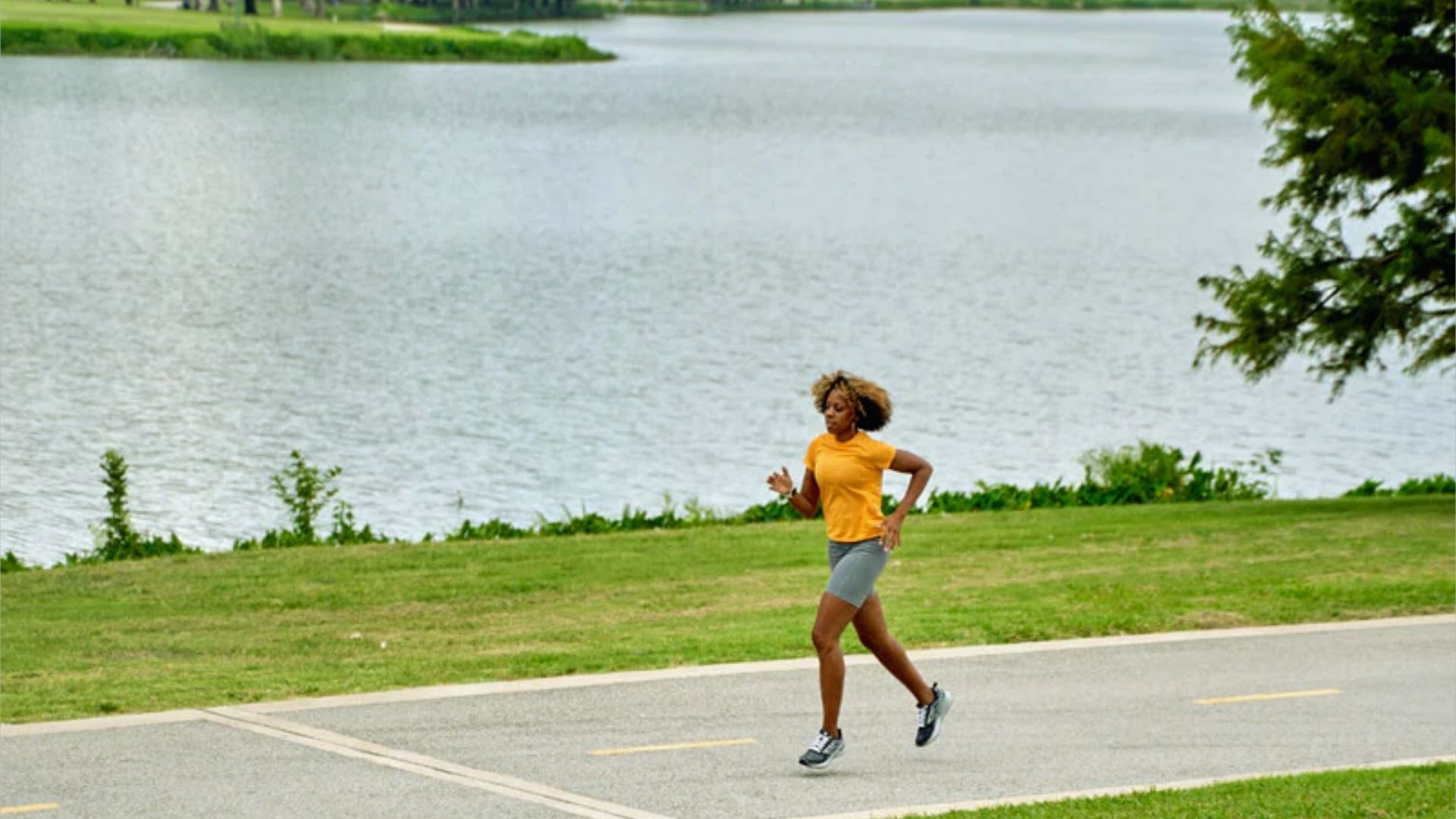 A person runs on a lakeside path, wearing a yellow shirt and gray shorts. The scene is bright and energetic, with greenery and calm water in the background.