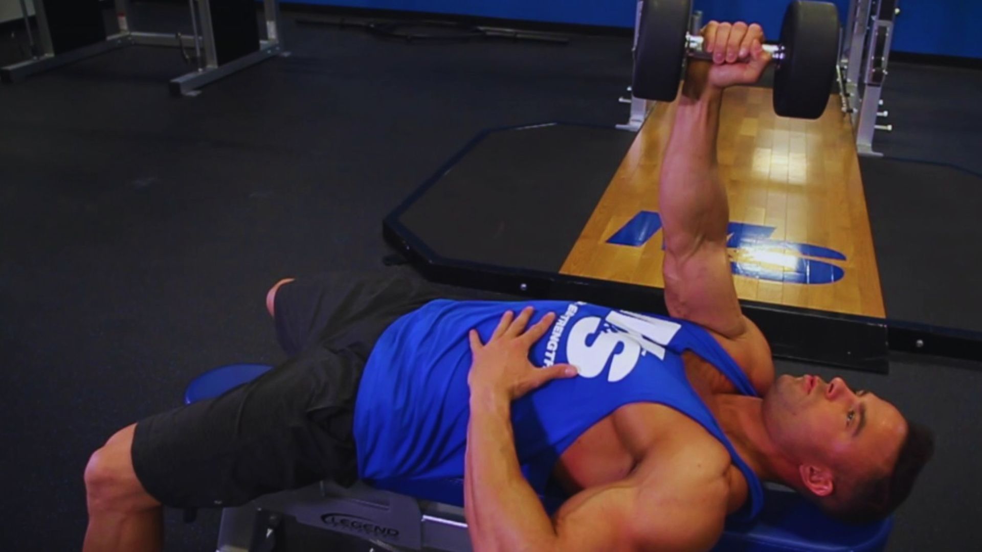A man in a blue tank top performs a dumbbell chest press on a bench in a gym, focusing intensely on his workout. The gym has a calm, focused atmosphere.