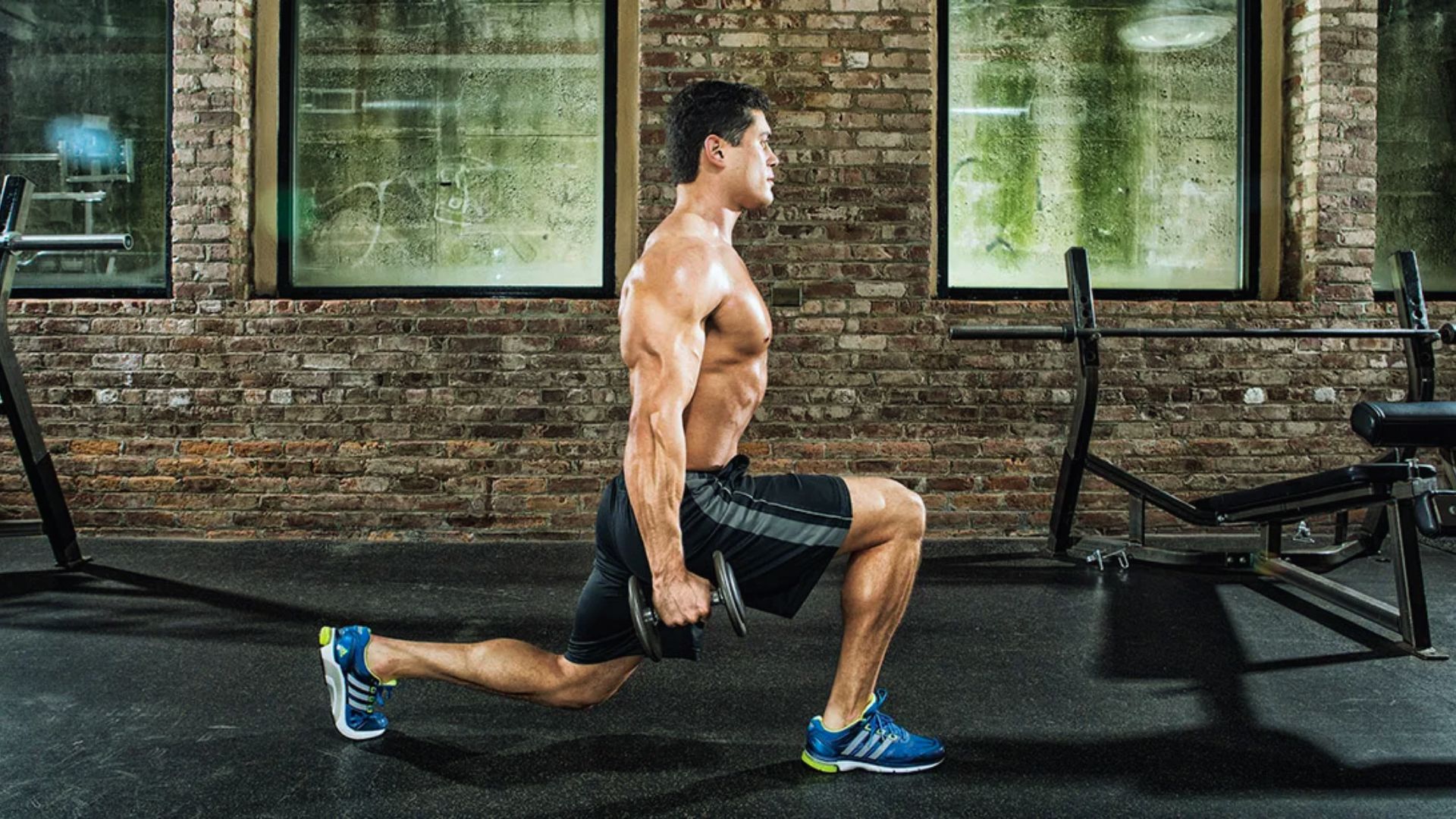 A muscular man performs a lunge with dumbbells in a gym. He's shirtless, wearing shorts and sneakers, emphasizing focus and strength in a brick-walled setting.