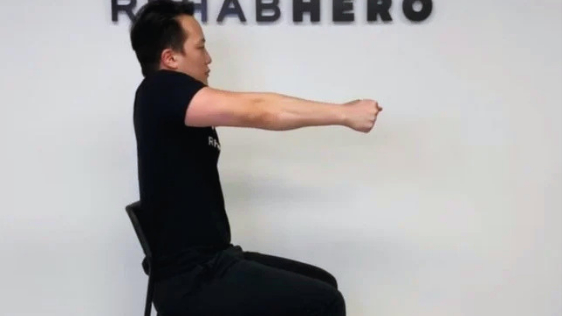 A person seated sideways performs a seated arm stretch. They extend their arms forward with a focused expression. Background reads "REHABHERO."