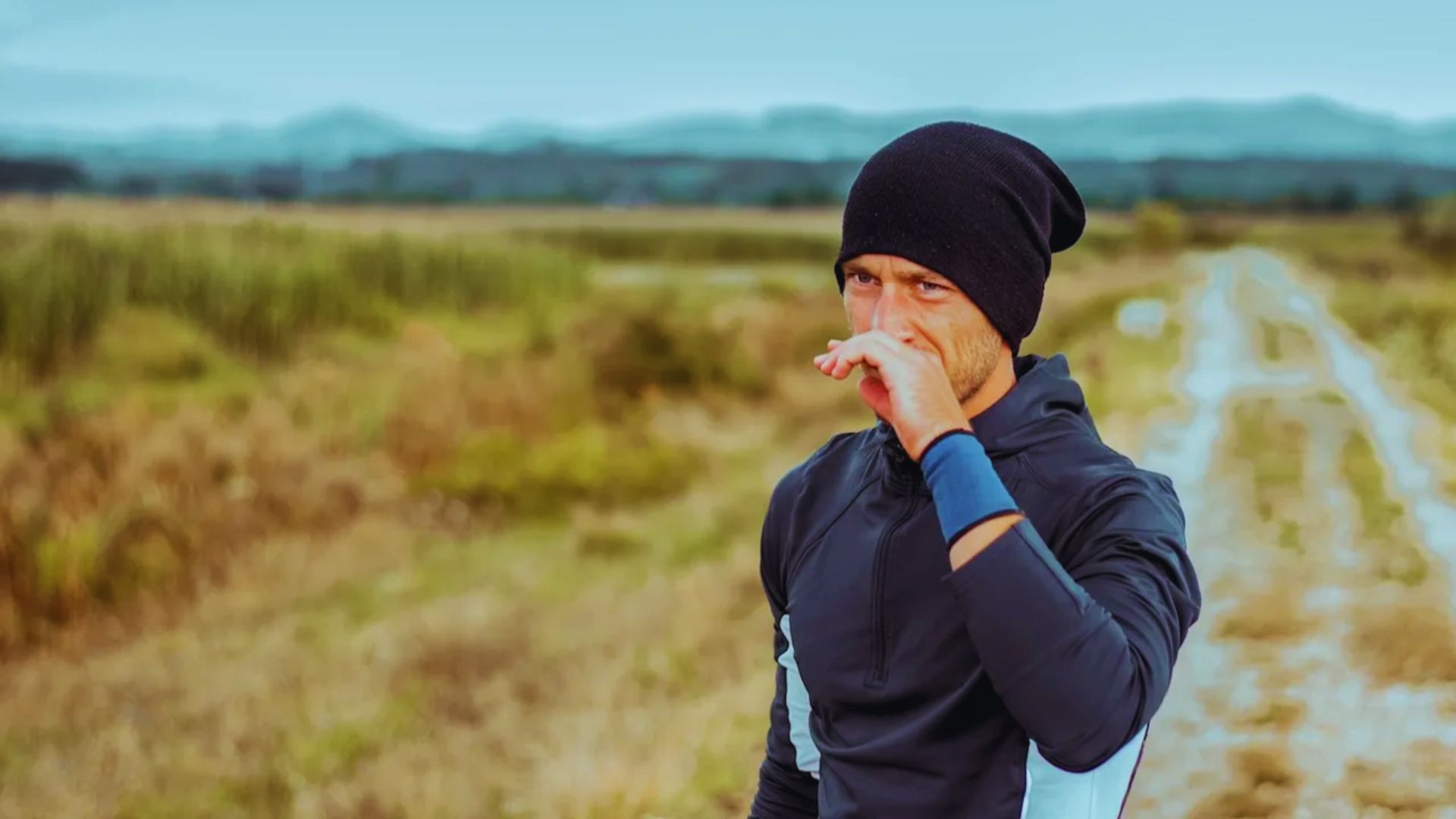Man in athletic wear and a beanie stands on a rural path, wiping his face. The landscape is a lush green meadow with distant hills. The mood is contemplative.