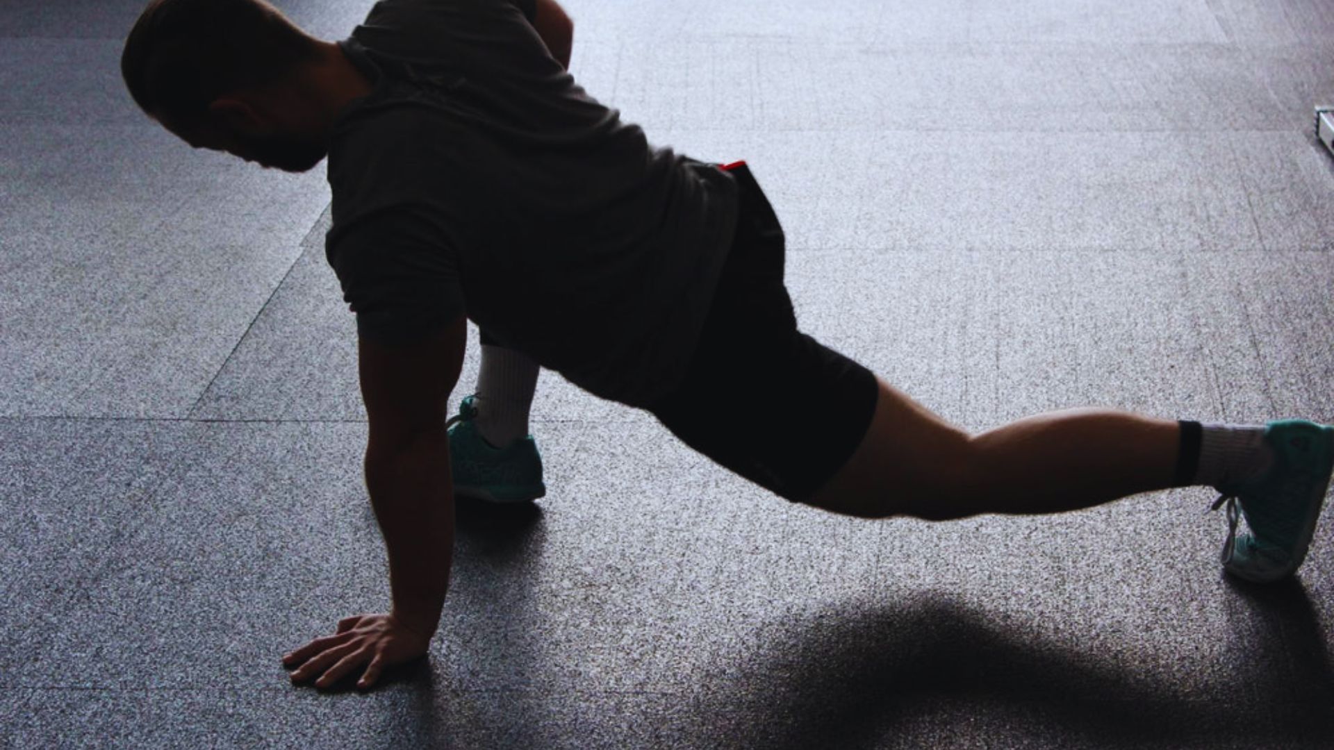 A person in athletic wear stretches on a gym floor, extending one leg back while supporting themselves with one hand. The scene is dimly lit, conveying focus.