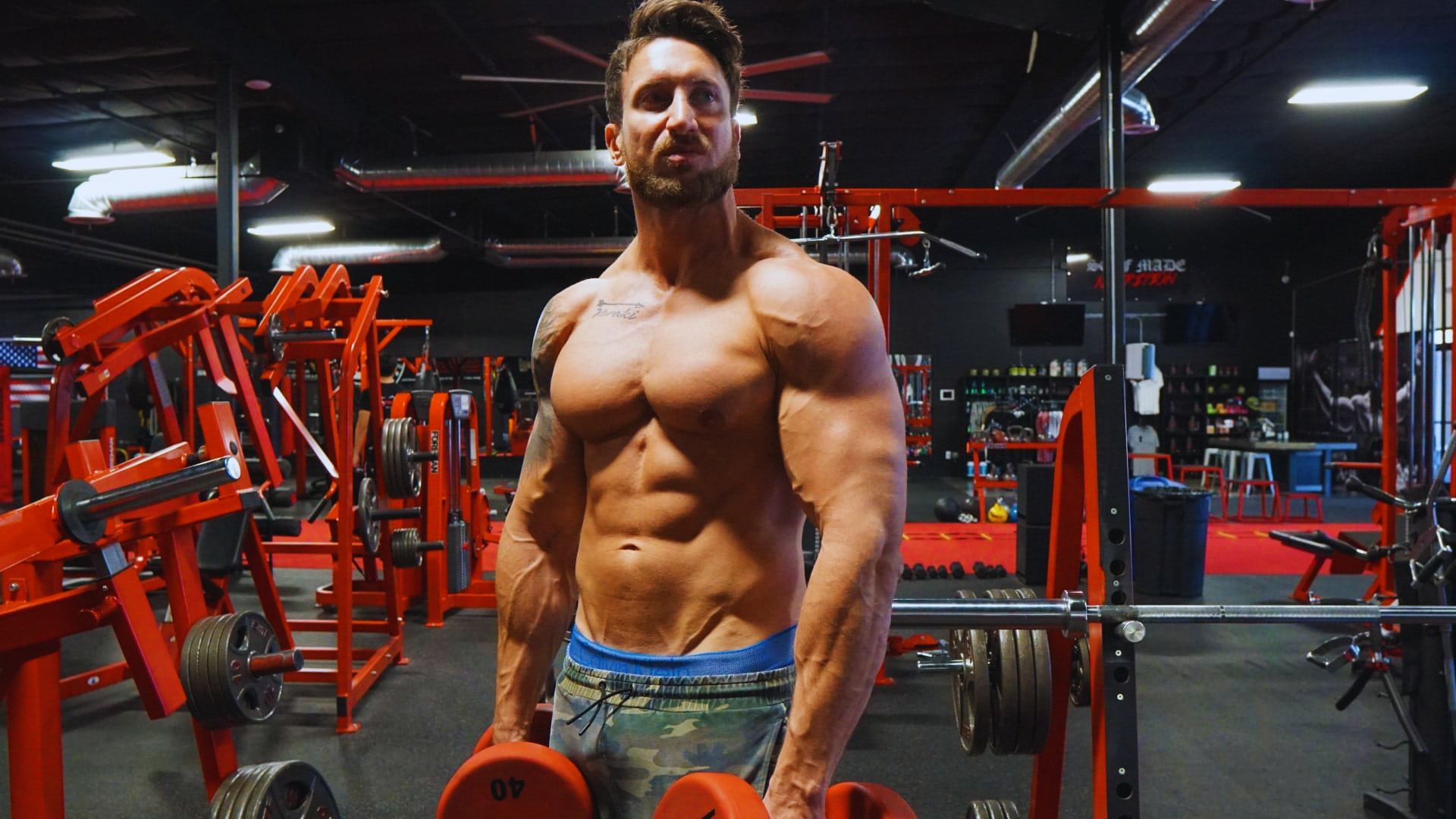 A shirtless man lifts weights in a gym, focusing on his workout amidst various gym equipment in the background.