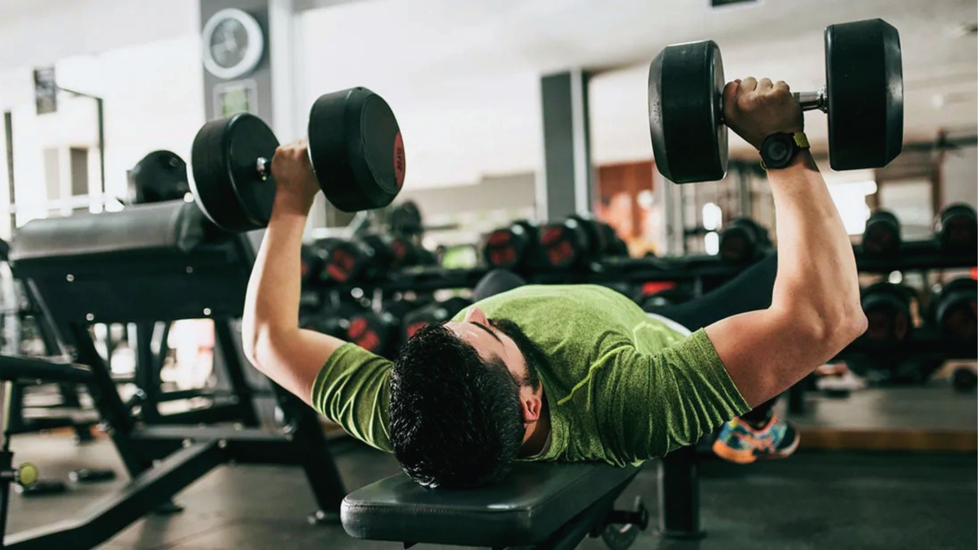 Man in a green shirt performs a dumbbell bench press at a gym. He is lying on a bench, surrounded by exercise equipment, focused and determined.
