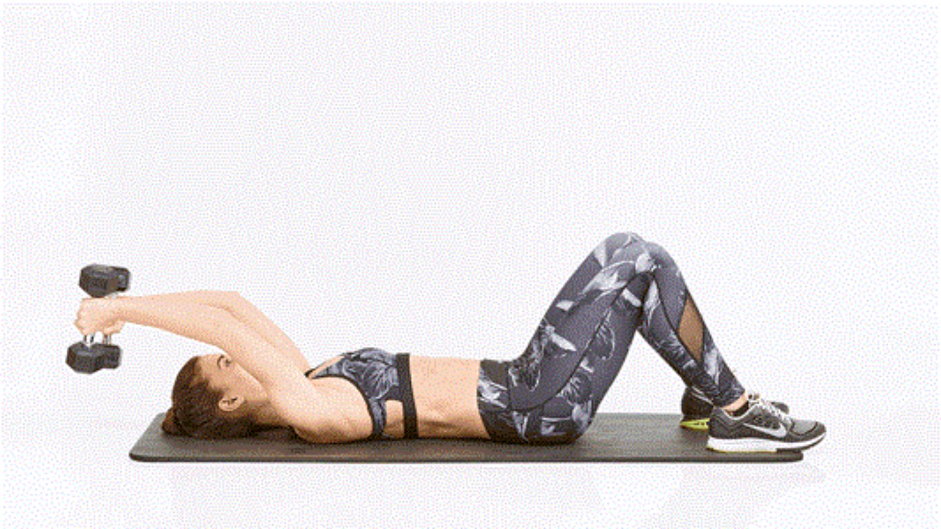 Woman lying on a yoga mat, performing a dumbbell floor press. Her knees are bent, feet flat, and arms extended, conveying focus and strength.