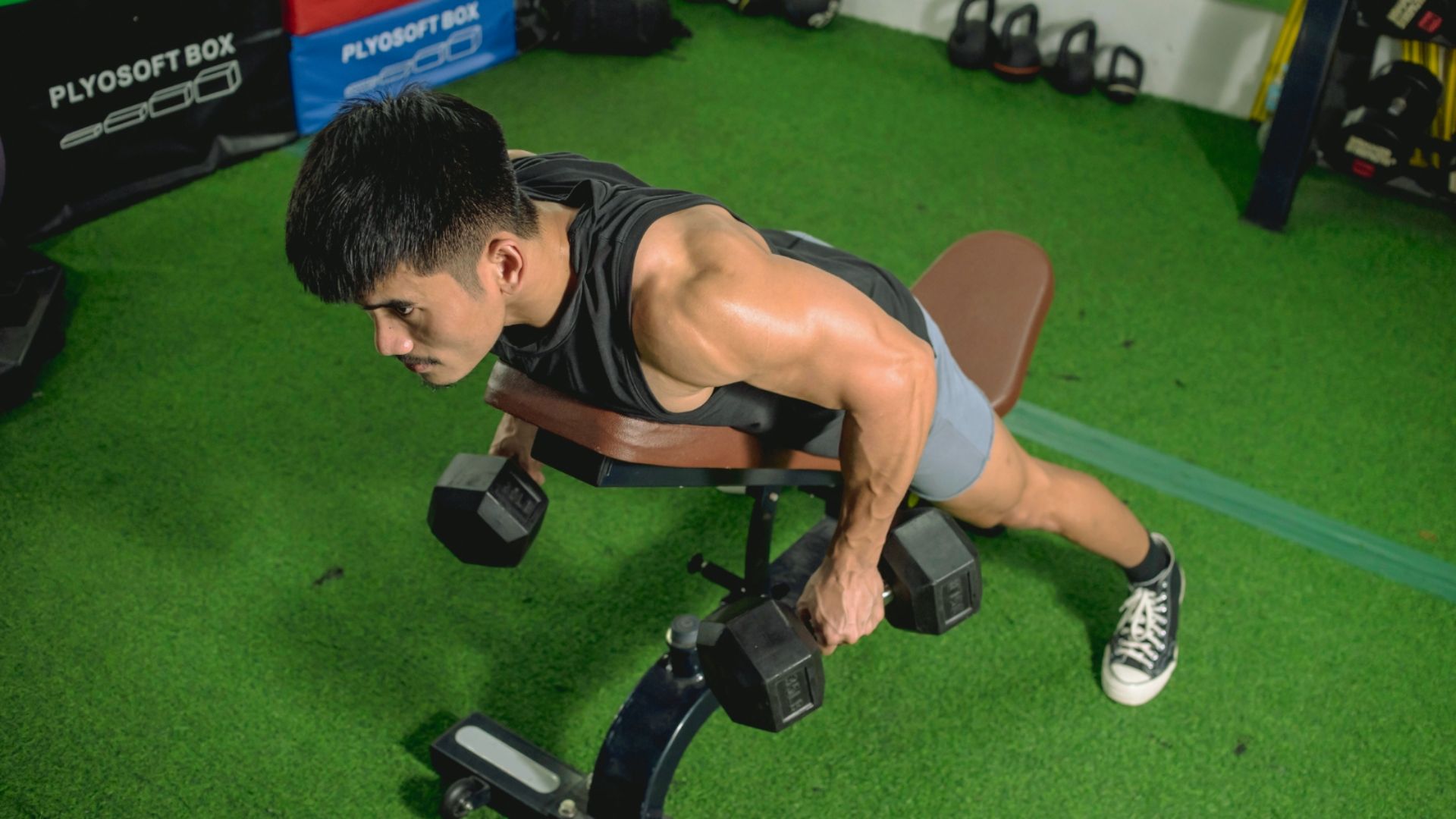A man in a gym performs a prone dumbbell row on a bench. He wears a black sleeveless shirt and shorts. The gym floor is covered in green turf.
