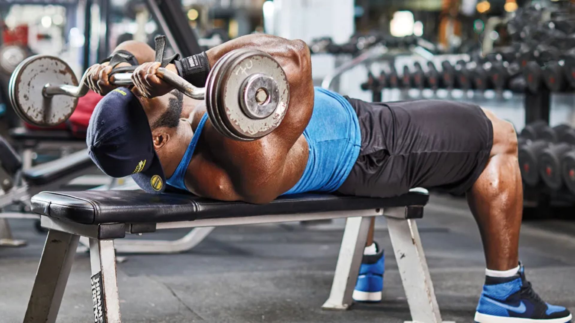 Man in blue shirt bench pressing a barbell in a gym. He is lying on a bench, focusing on lifting weights."