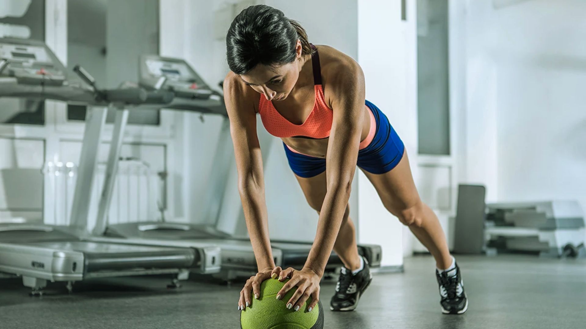 A woman in athletic wear performs a plank on a medicine ball in a gym, conveying focus and determination. Treadmills are visible in the background.
