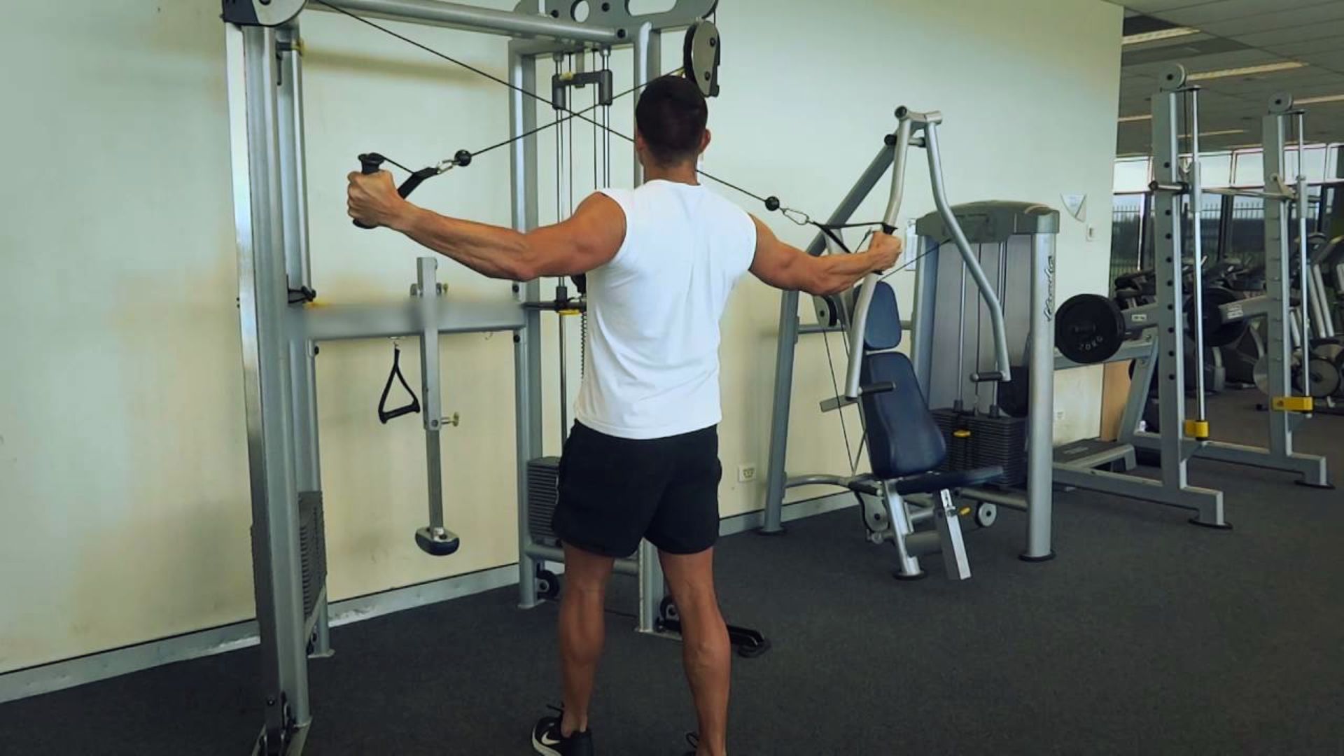 Man in a gym wearing a white tank top and black shorts performs a cable crossover exercise with focused intensity. Machines and weights in the background.