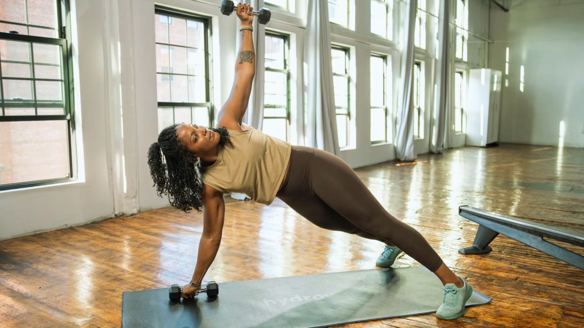 Woman in gym performs side plank with dumbbells on yoga mat, sunlight streams through large windows, creating an energetic, focused atmosphere.