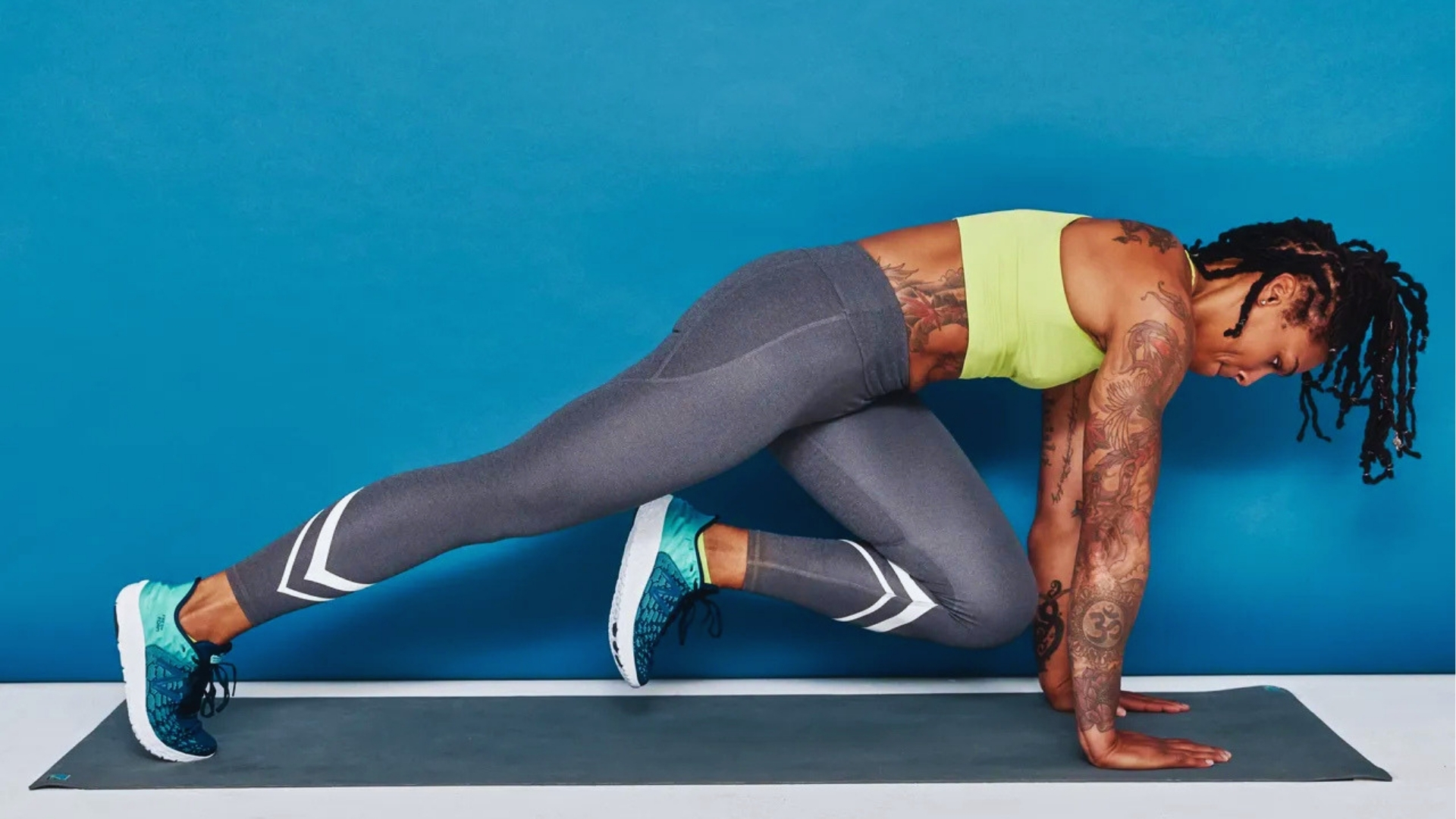 A person with tattoos is doing a mountain climber exercise on a yoga mat. They're wearing a lime green top, gray leggings, and teal sneakers.