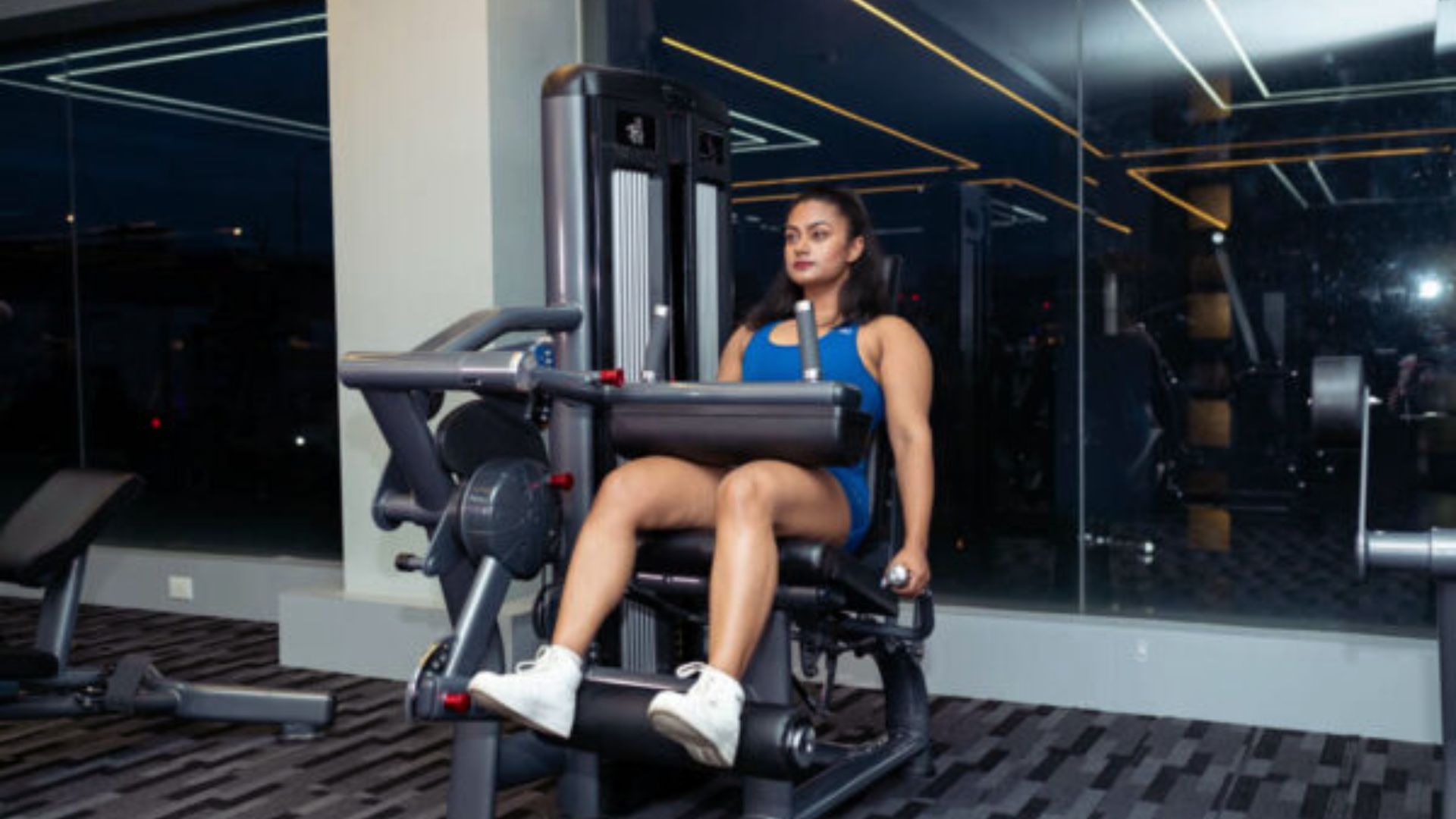 A woman in a blue workout outfit uses a leg press machine in a modern gym with dark flooring and mirrored walls, looking focused and determined.