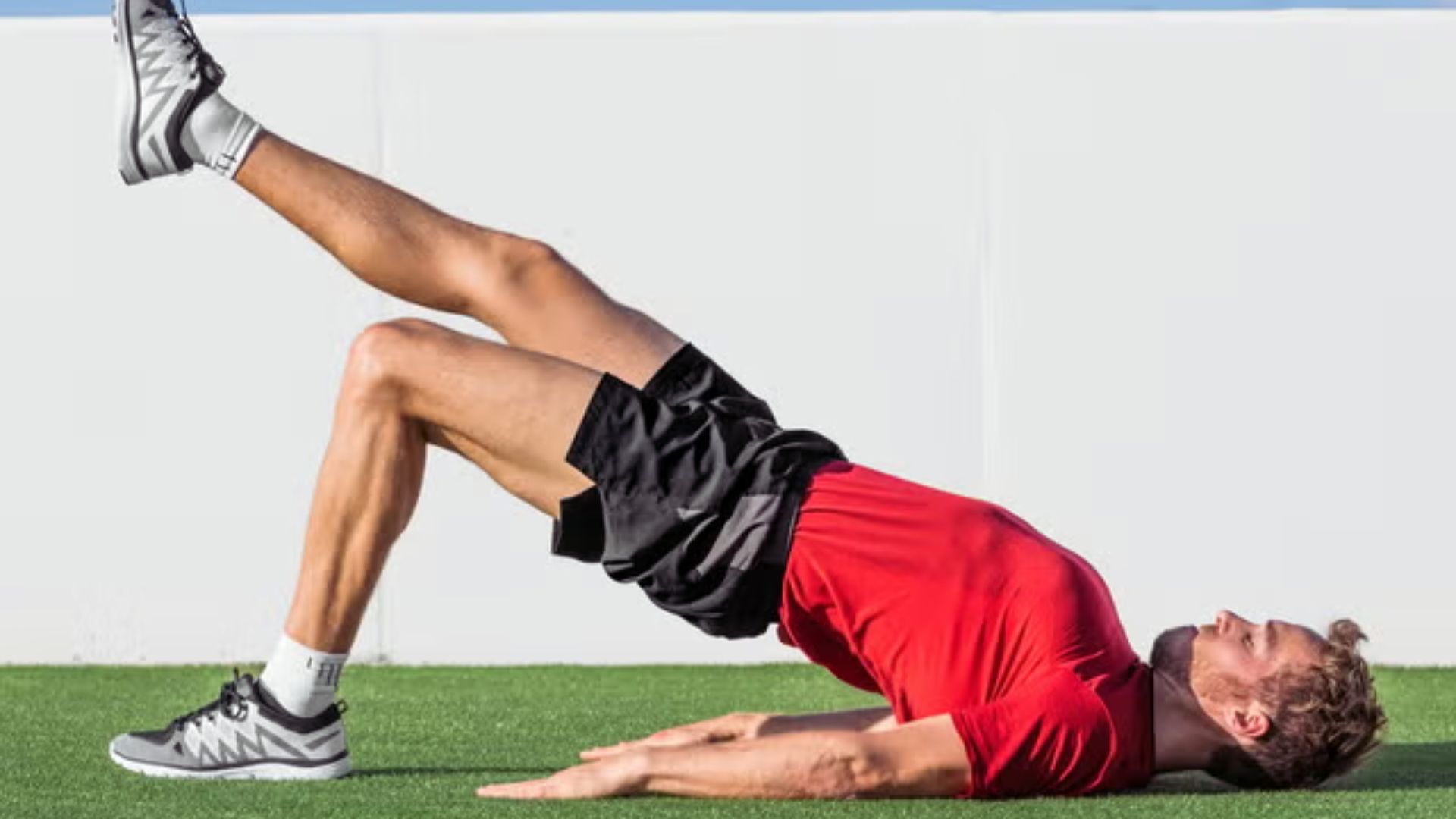 Man in a red shirt and black shorts performs a single-leg bridge exercise on grass. One leg is raised, showcasing balance and strength.