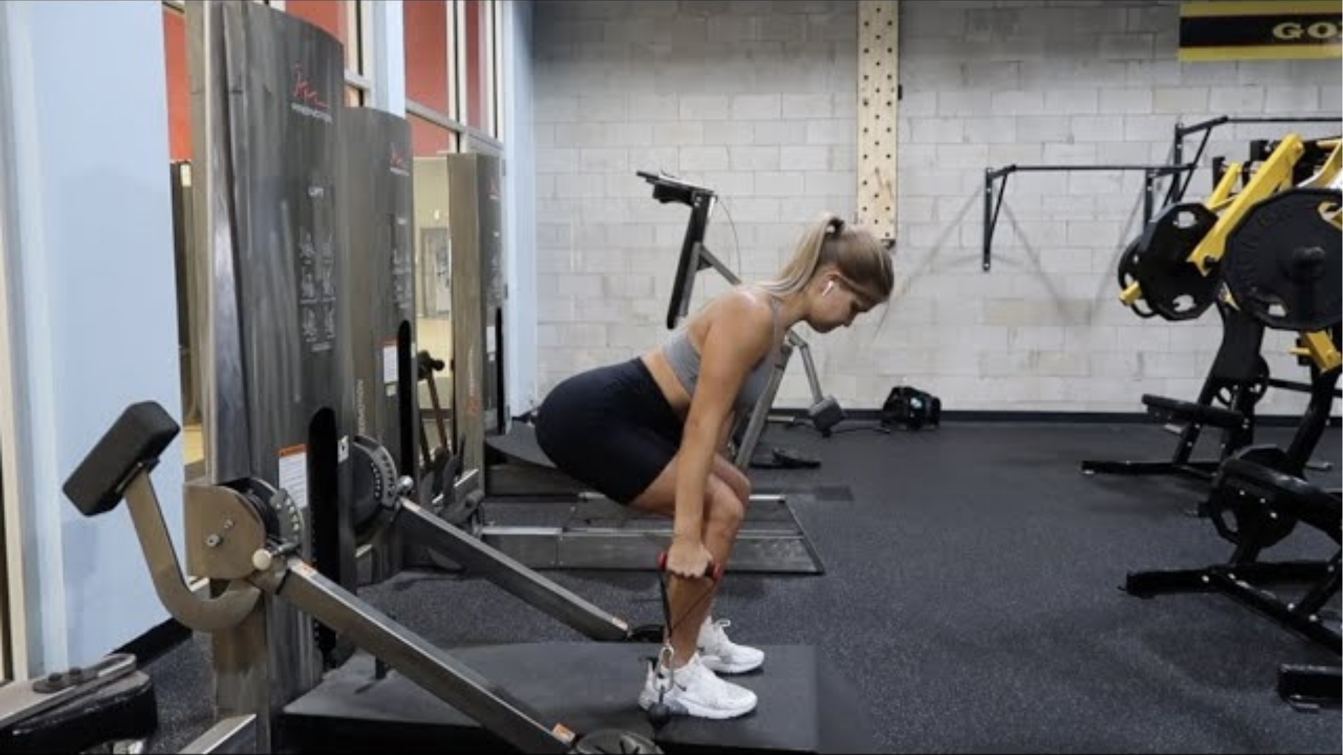 A woman exercises in a gym using a cable row machine. She wears athletic gear, and the room is equipped with various fitness machines, conveying focus and determination.