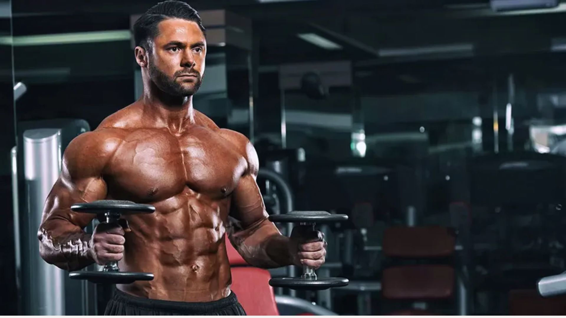 Muscular man intensely lifts dumbbells in a dimly lit gym, showcasing strong physique and focused expression, surrounded by gym equipment.