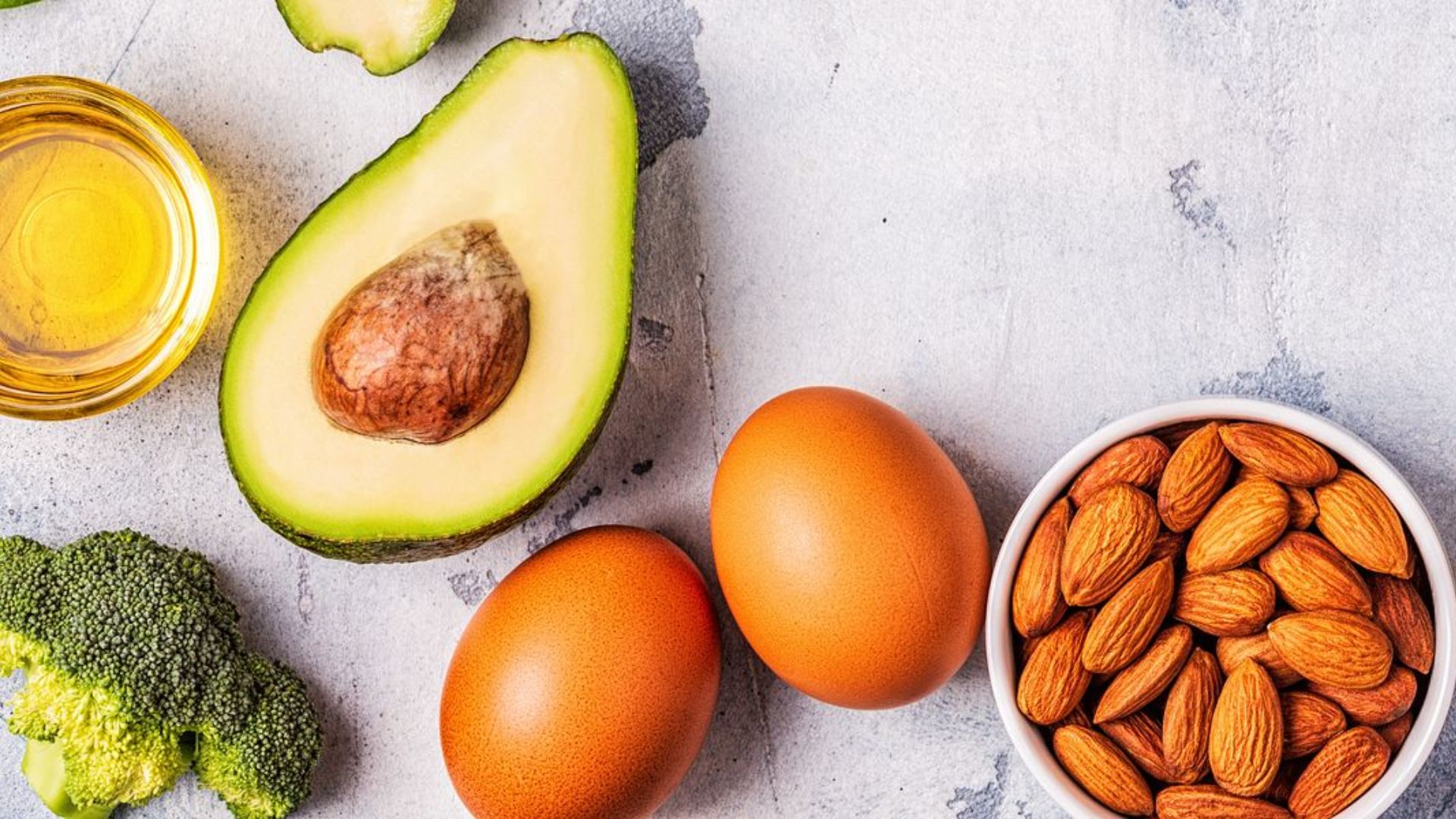 A flat lay of healthy foods on a light surface: halved avocado, bowl of almonds, two eggs, broccoli, and a small dish of golden oil, conveying freshness.