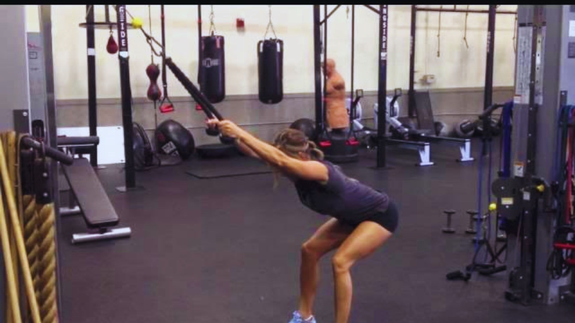 A woman in athletic wear performs a stretching exercise using TRX straps in a gym. The setting is equipped with punching bags and workout equipment.