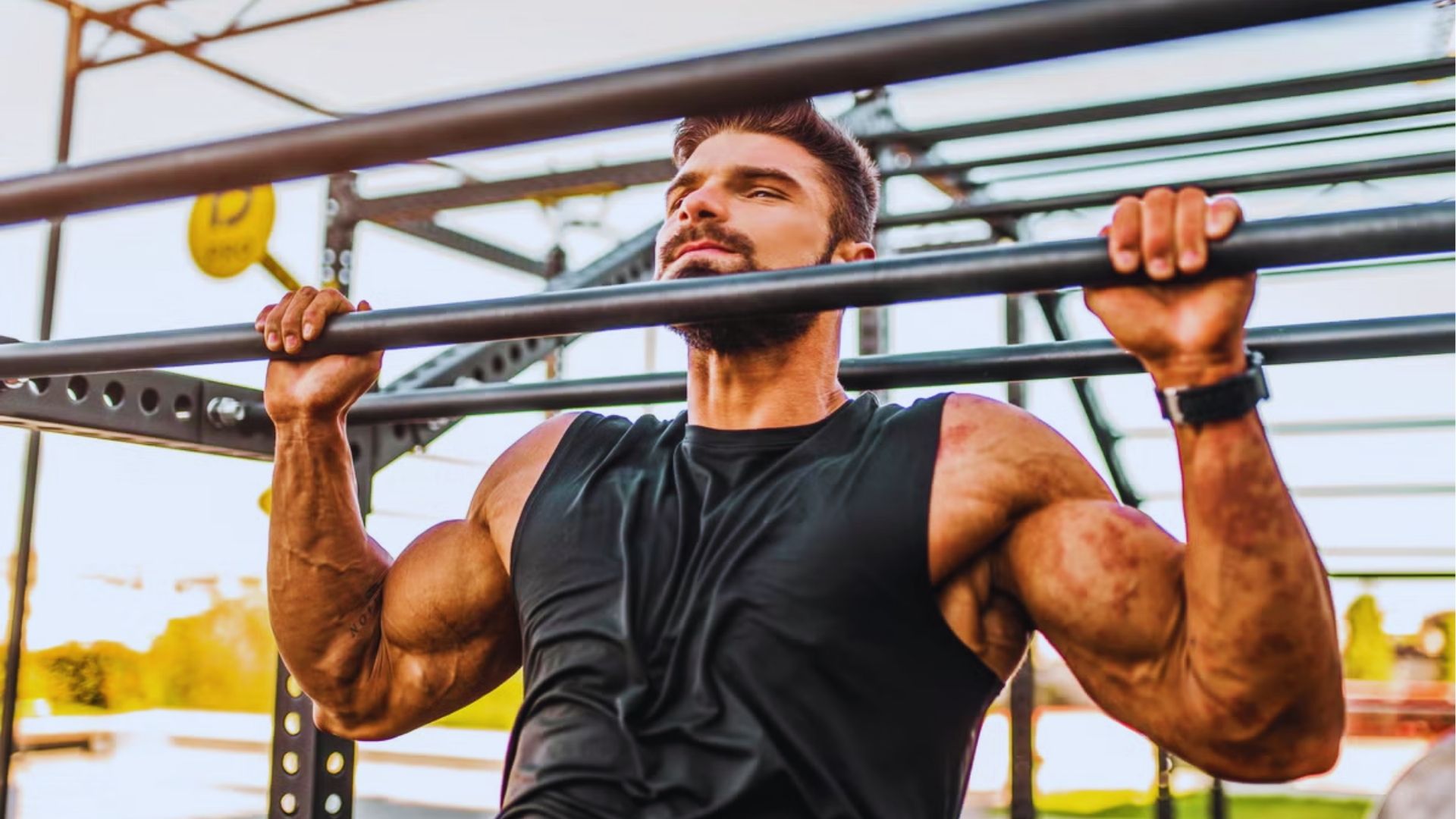 A muscular man in a black tank top performs a pull-up on an outdoor metal bar. The background shows a bright, sunny day, conveying strength and focus.