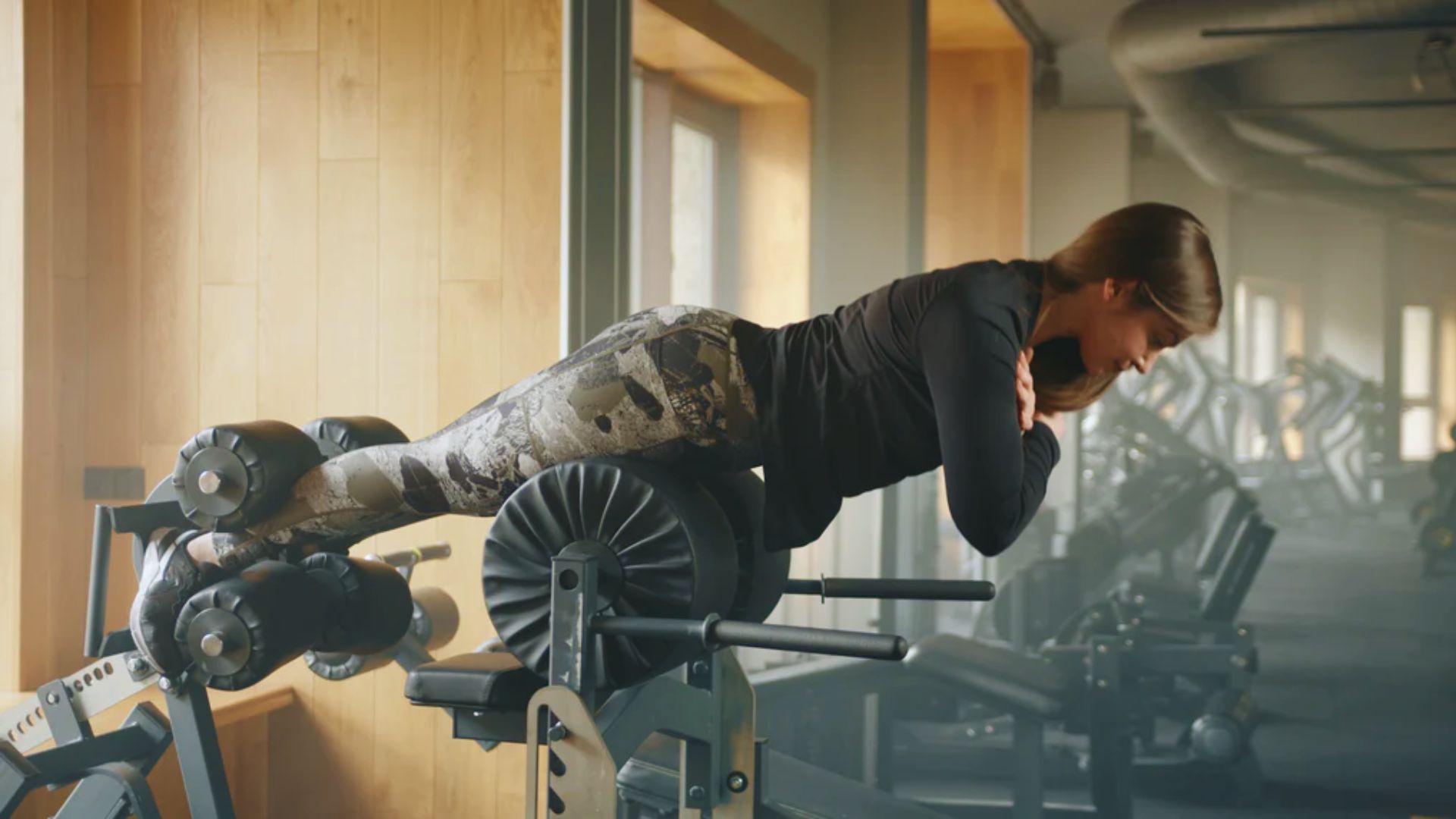 A woman in workout attire uses a hyperextension bench in a gym, focusing intently. The room is spacious, with exercise equipment in the background.