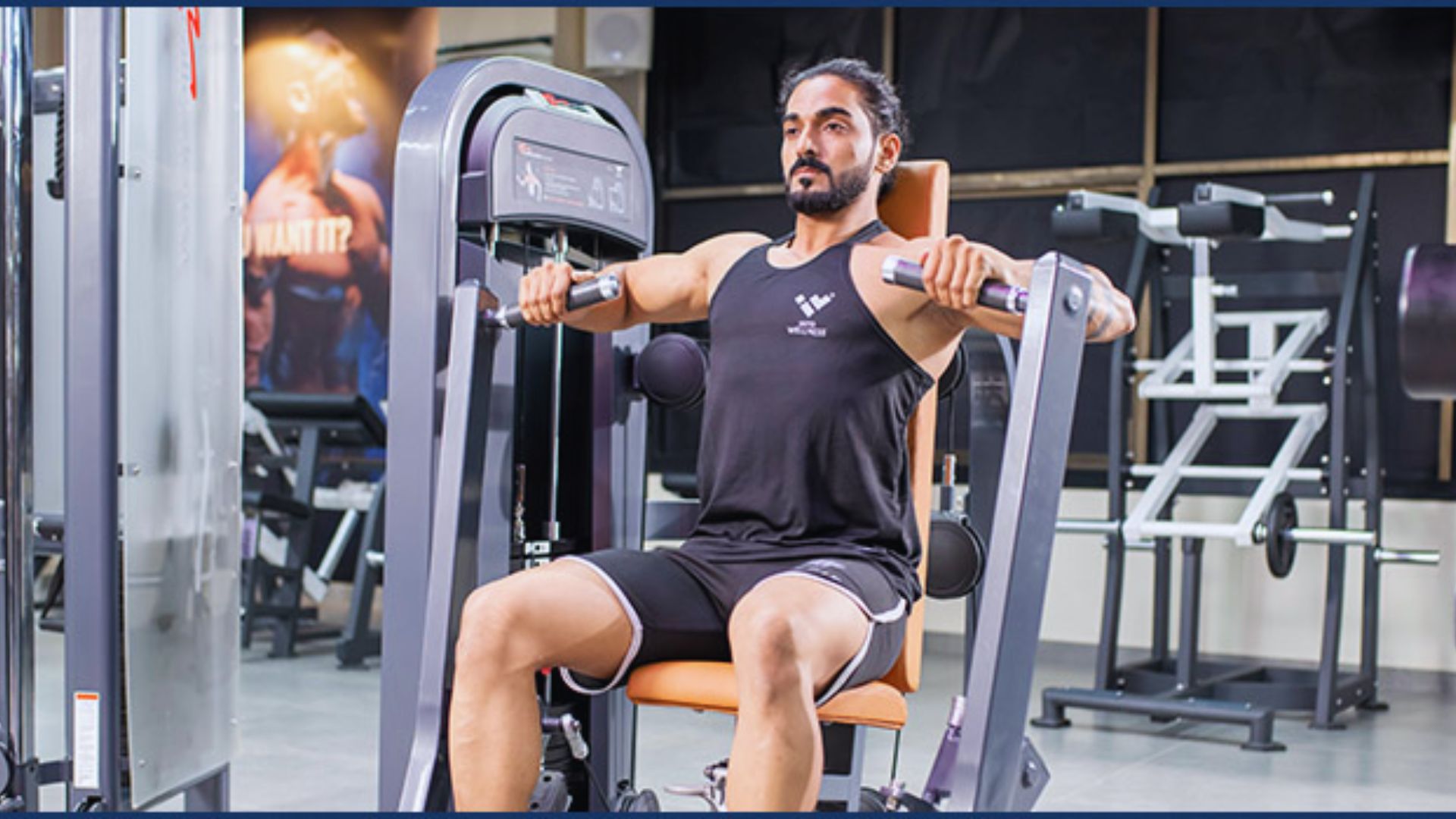 A man with a beard is focused and seated on a shoulder press machine in a gym, wearing a black tank top and shorts. Equipment is visible in the background.