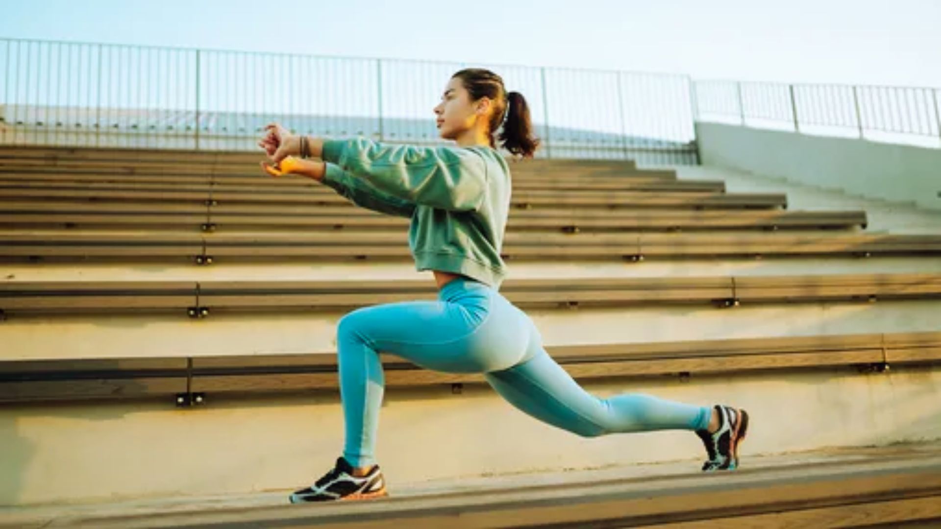 A woman in athletic wear performs a lunge on outdoor bleachers. She's focused and determined, with a clear sky in the background, evoking energy and fitness.