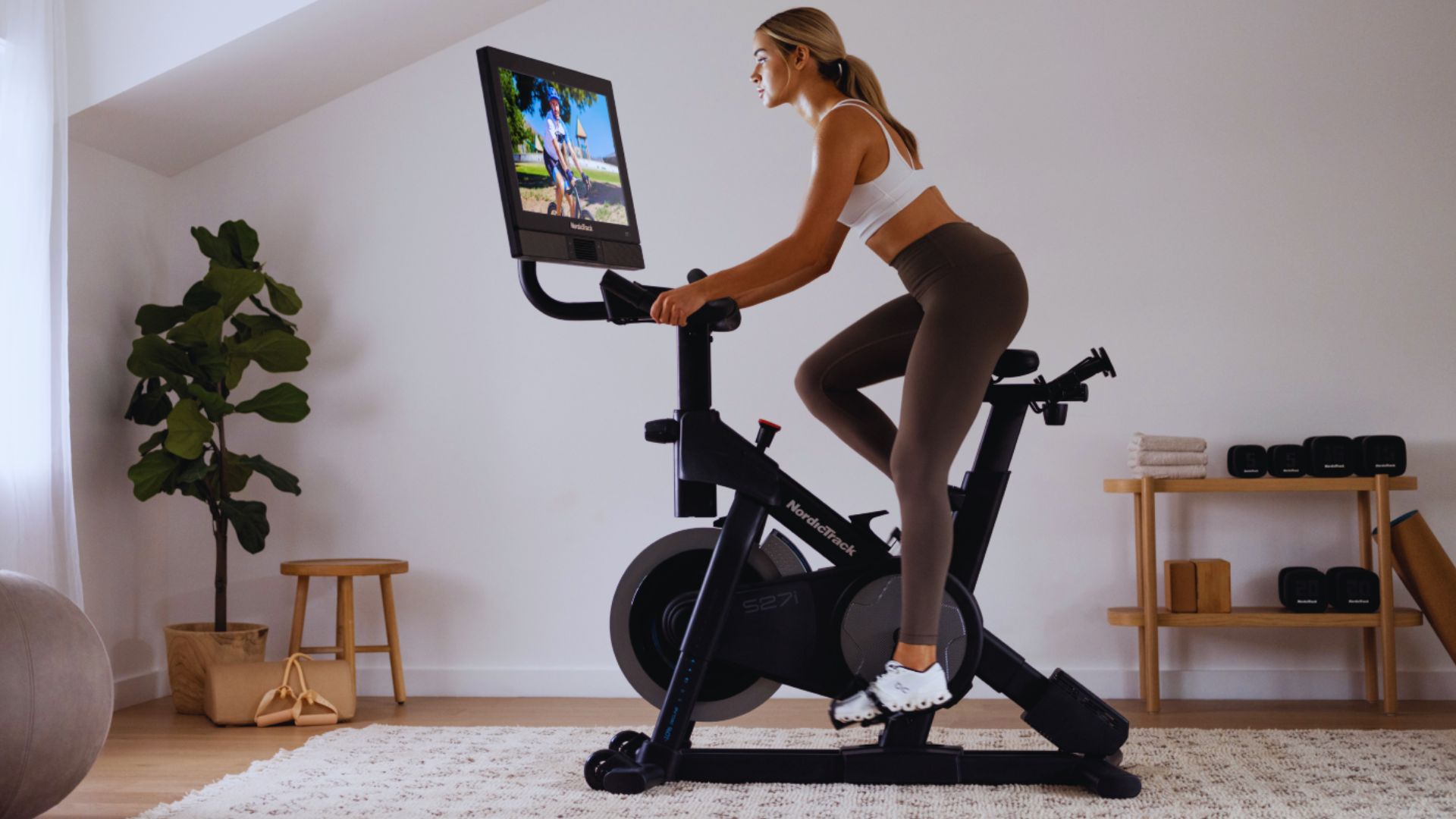 A woman in workout clothes rides a stationary bike with a screen displaying a scenic route. The room is minimalistic, featuring plants and light decor.