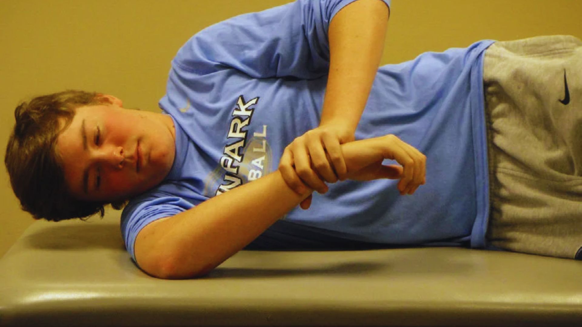 A young person in a blue shirt and gray pants lies on their side on a therapy table, focusing on wrist exercises. The mood is calm and focused.
