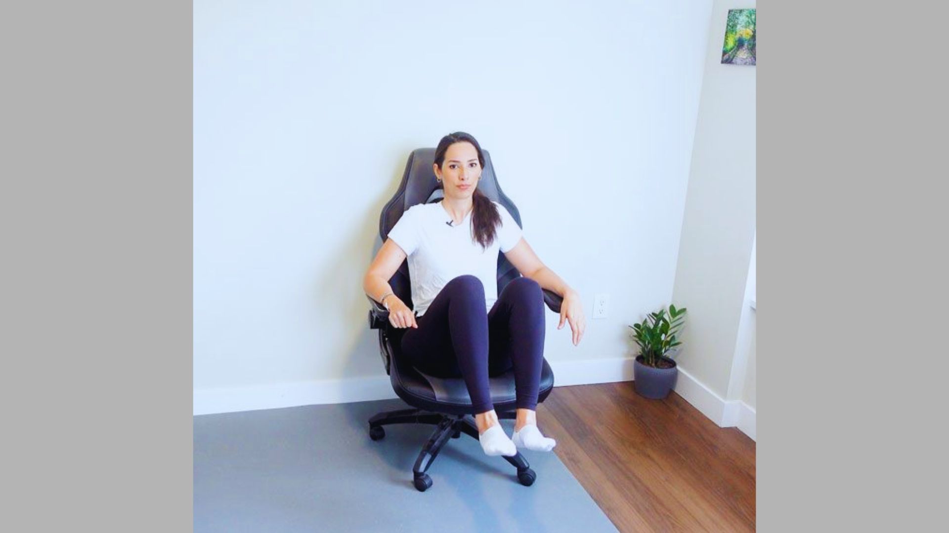 A person in a white shirt and black leggings performs a seated exercise on an office chair in a bright room with a plant in the corner, conveying focus.