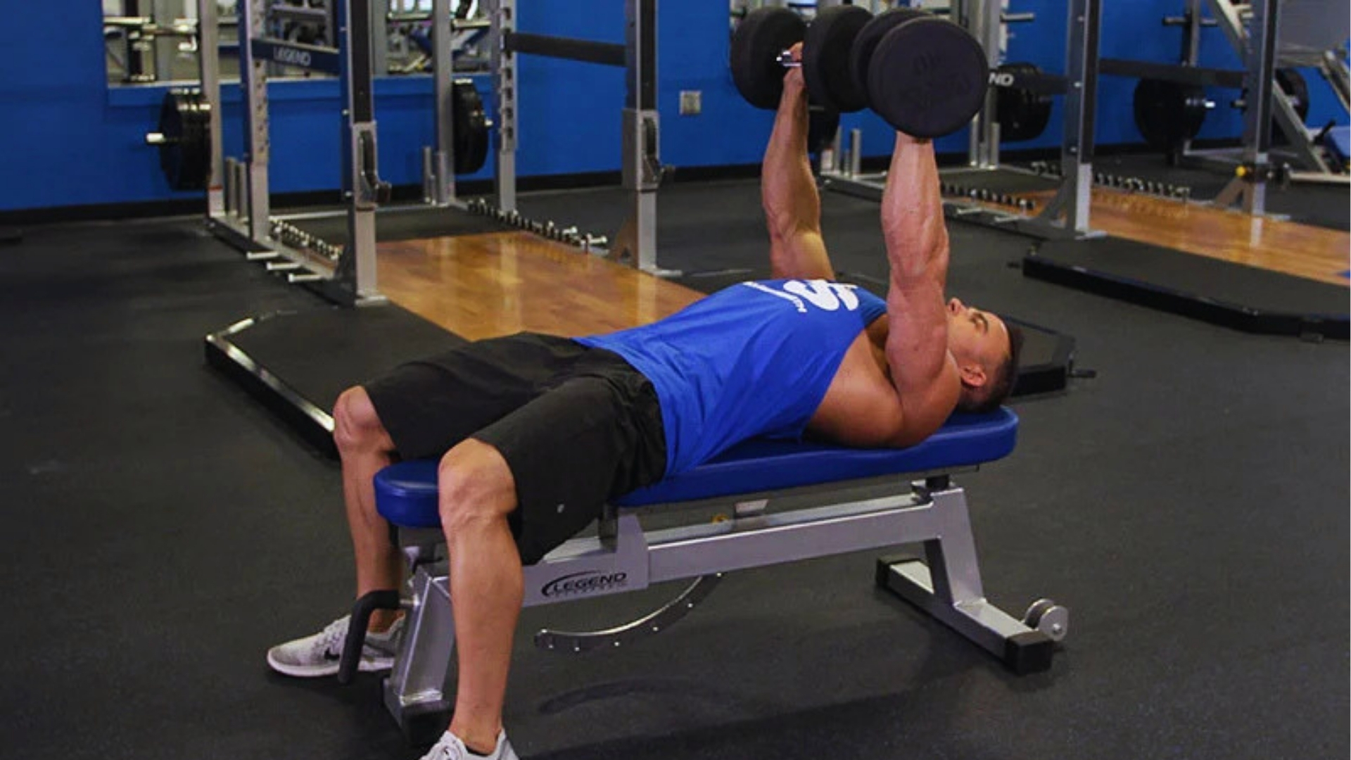 Man in a blue tank top performing a bench press with dumbbells in a gym. He is lying on a bench, focused and engaged, with exercise equipment around.