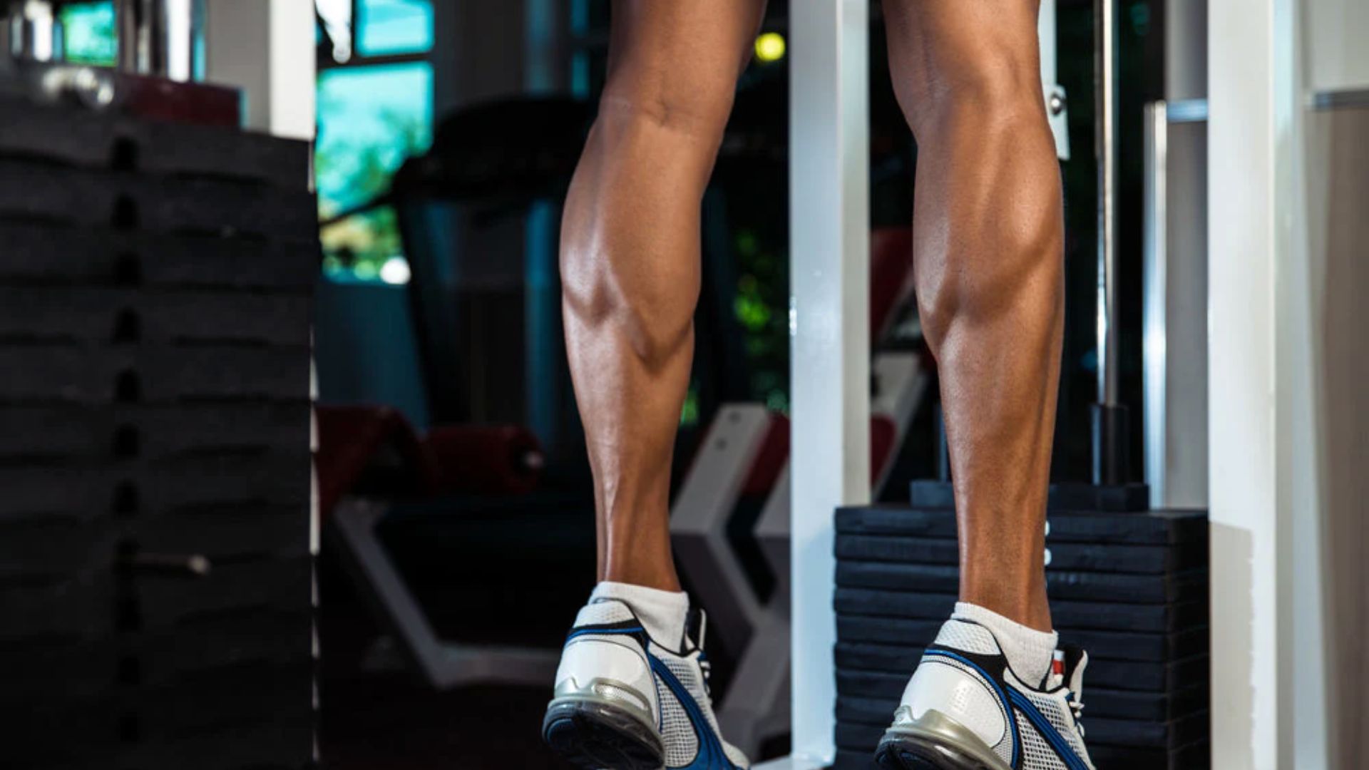 Person's muscular calves in athletic shoes performing a calf raise at the gym, with weight stacks and gym equipment blurred in the background.