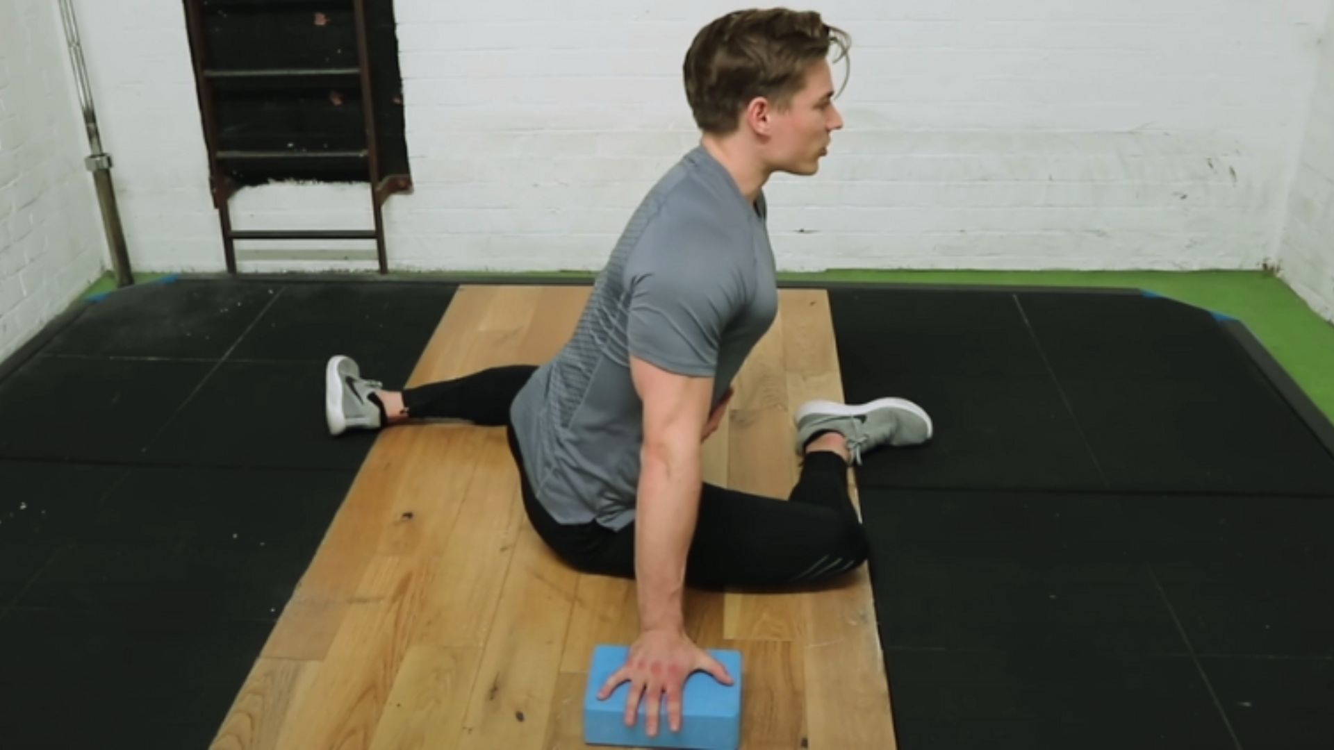 A person in a gray t-shirt and black pants performs a pigeon pose stretch on a wooden floor. They use a blue foam block for support, displaying focus and balance.