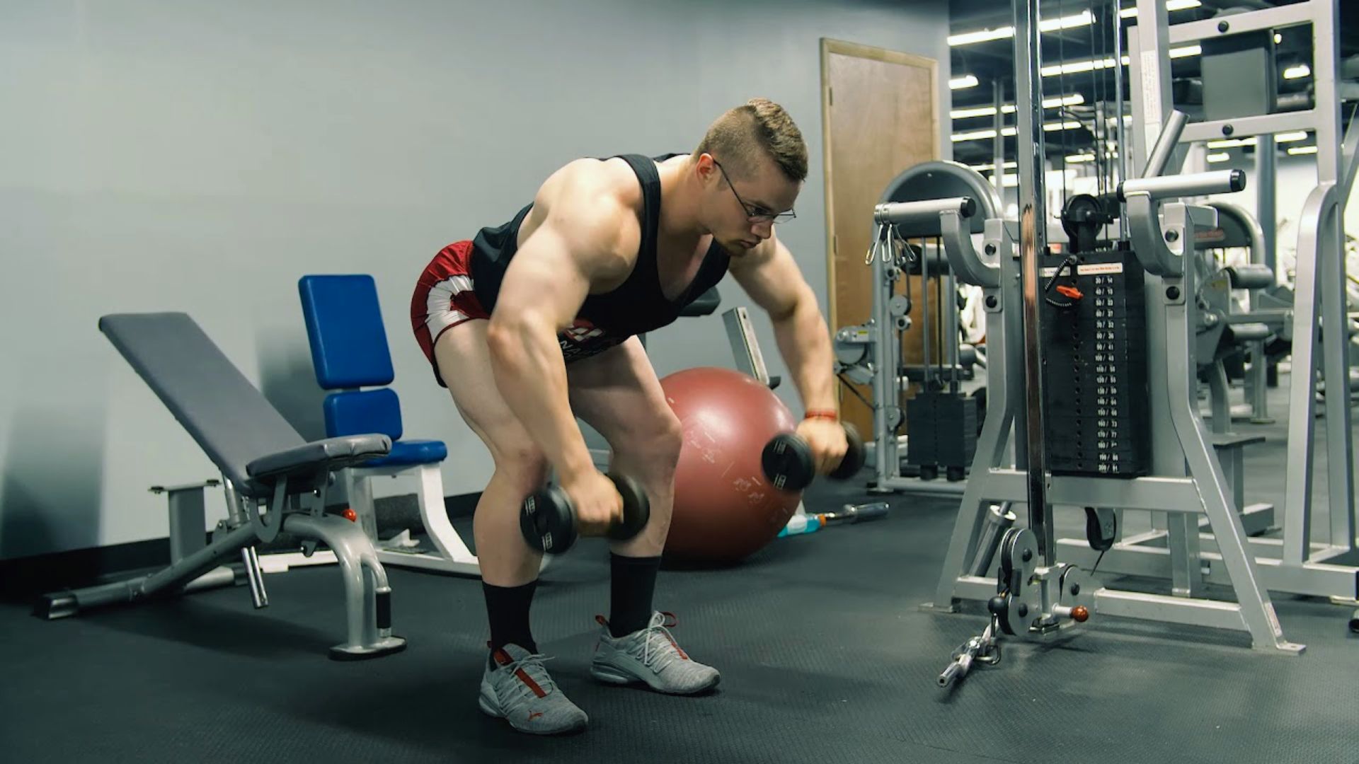 A person in a gym performs a bent-over dumbbell exercise. They wear a tank top and shorts, surrounded by fitness equipment, creating a focused atmosphere.