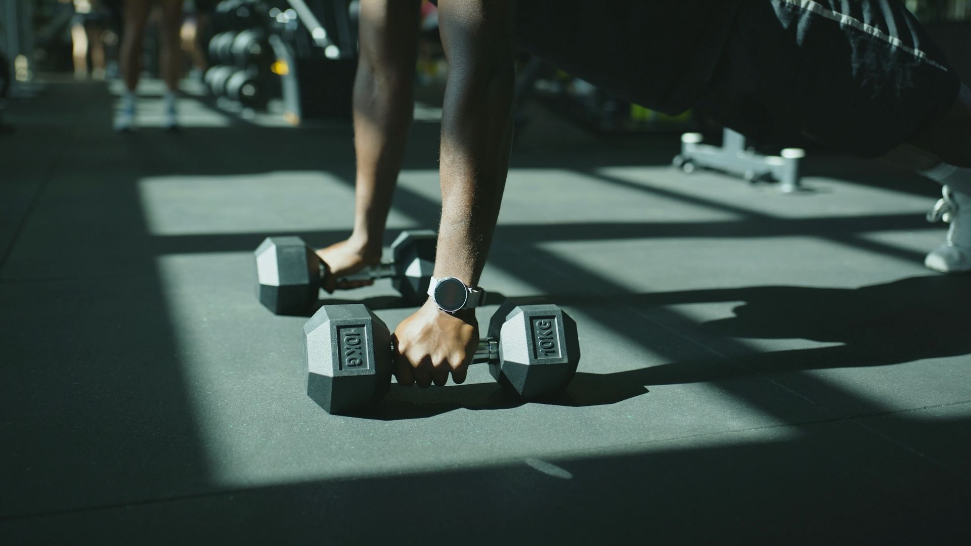 A person performs a plank using dumbbells in a sunlit gym. The focus is on their hands and arms, conveying strength and determination.