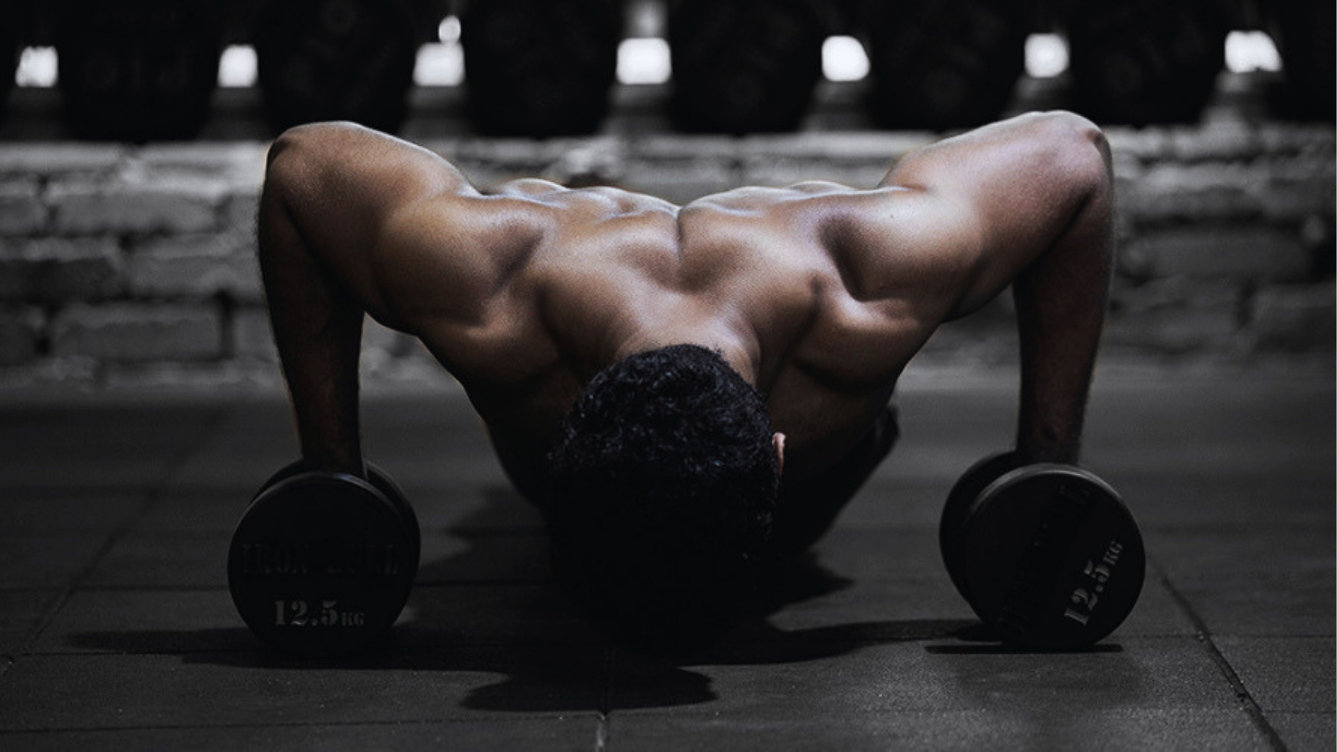 A muscular person performs a push-up on dumbbells in a dimly lit gym. Strong back muscles are highlighted, conveying strength and focus.