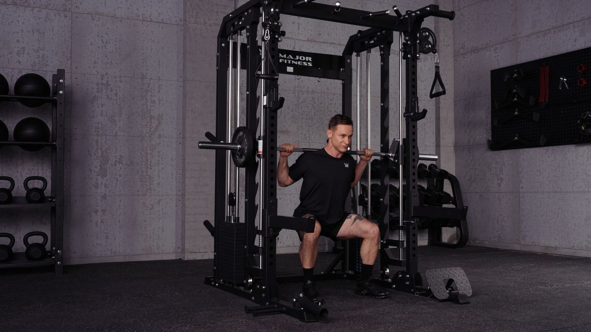 A man in a black T-shirt and shorts performs squats with a barbell on a Smith machine in a modern gym. Kettlebells and equipment are visible in the background.