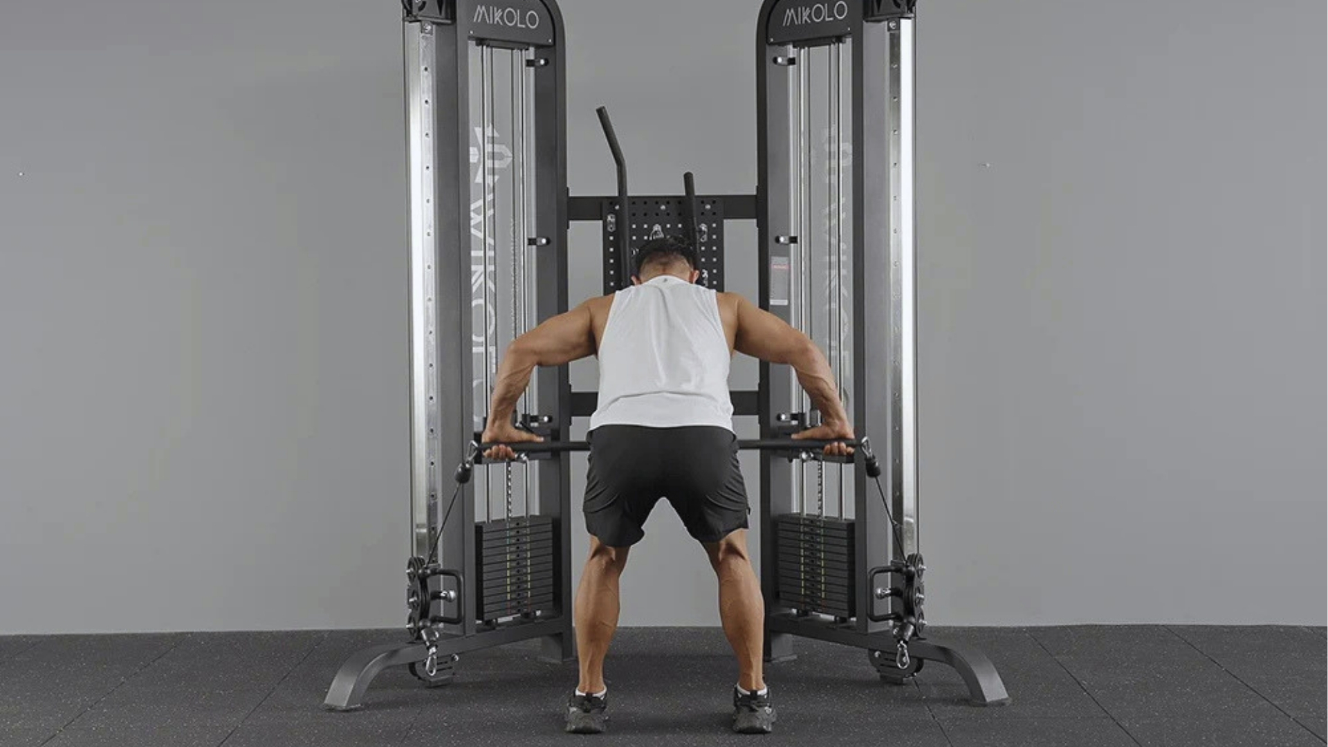 A man in a white tank top and black shorts uses a cable machine in a gym, standing with legs apart, focused and exerting effort.