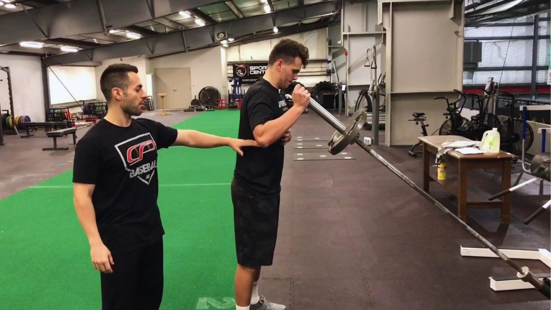 Two men in a gym; one is performing a landmine press with a barbell while the other assists, checking posture. The gym has green turf flooring.