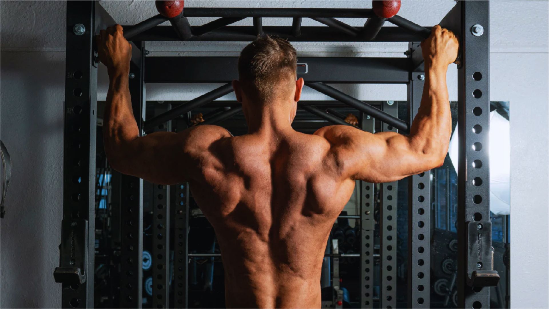 Muscular man performing a pull-up on a gym bar, showcasing defined back and shoulder muscles. The setting is a weightlifting gym, emphasizing strength and athleticism.